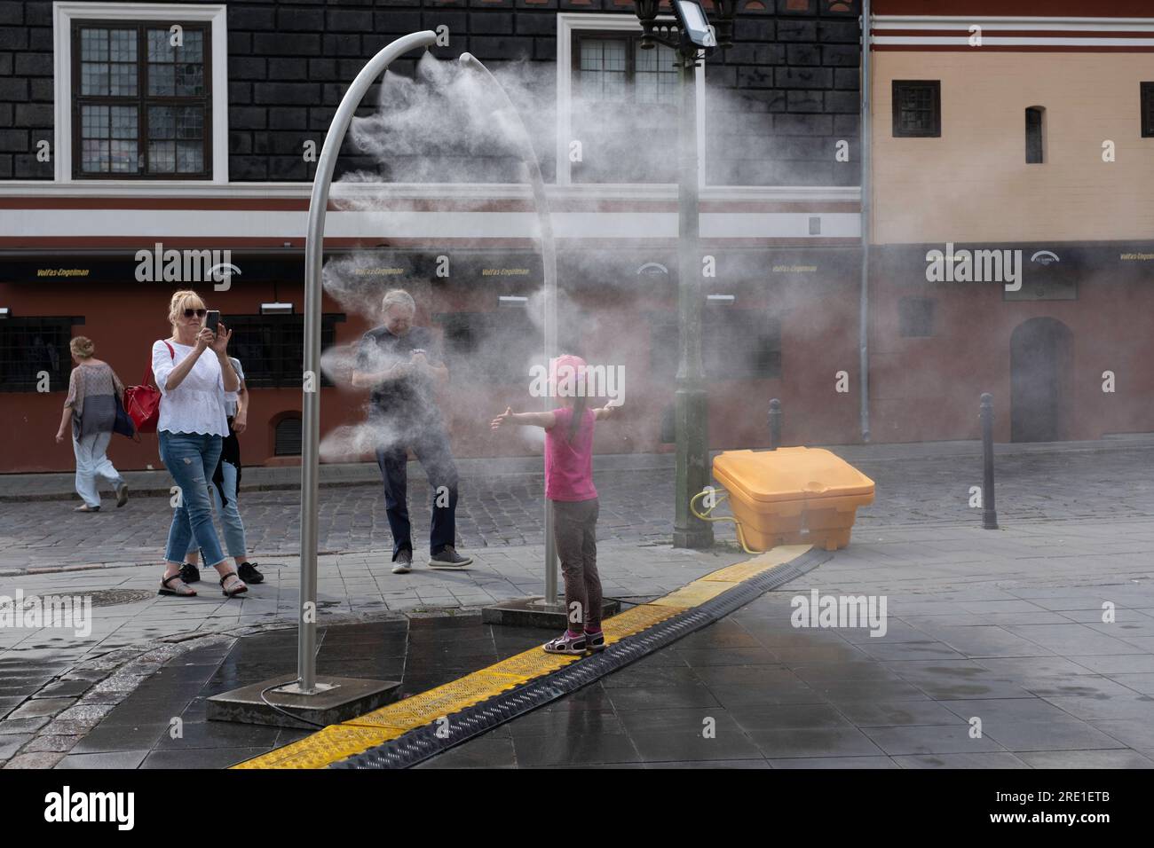 La ragazza viene raffreddata da un'installazione di spruzzi d'acqua su una piazza a Kaunas. I suoi genitori la fotografano. Pericolo di contaminazione da Legionella Foto Stock