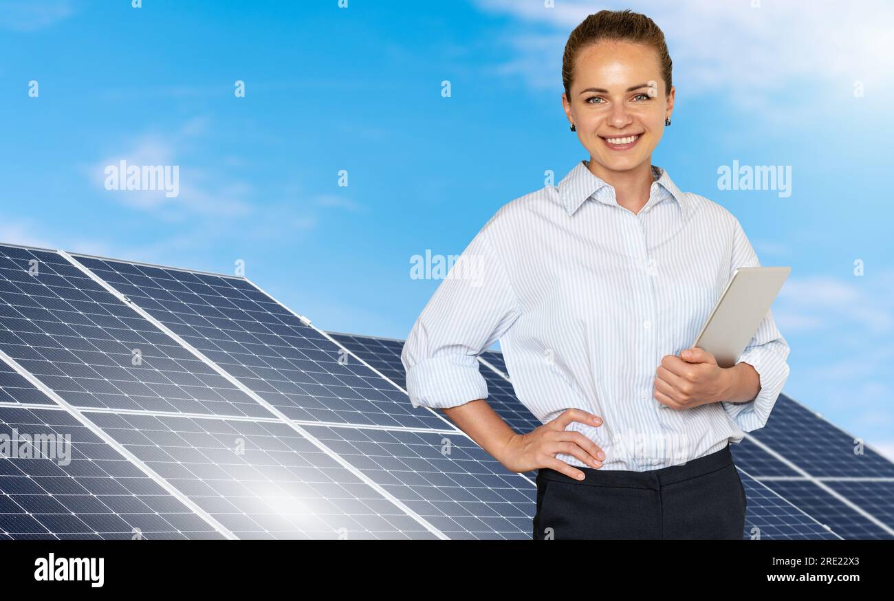 Giovane ingegnere in piedi sullo sfondo di una fattoria di energia solare che tiene in mano un tablet digitale e sorride. Foto Stock