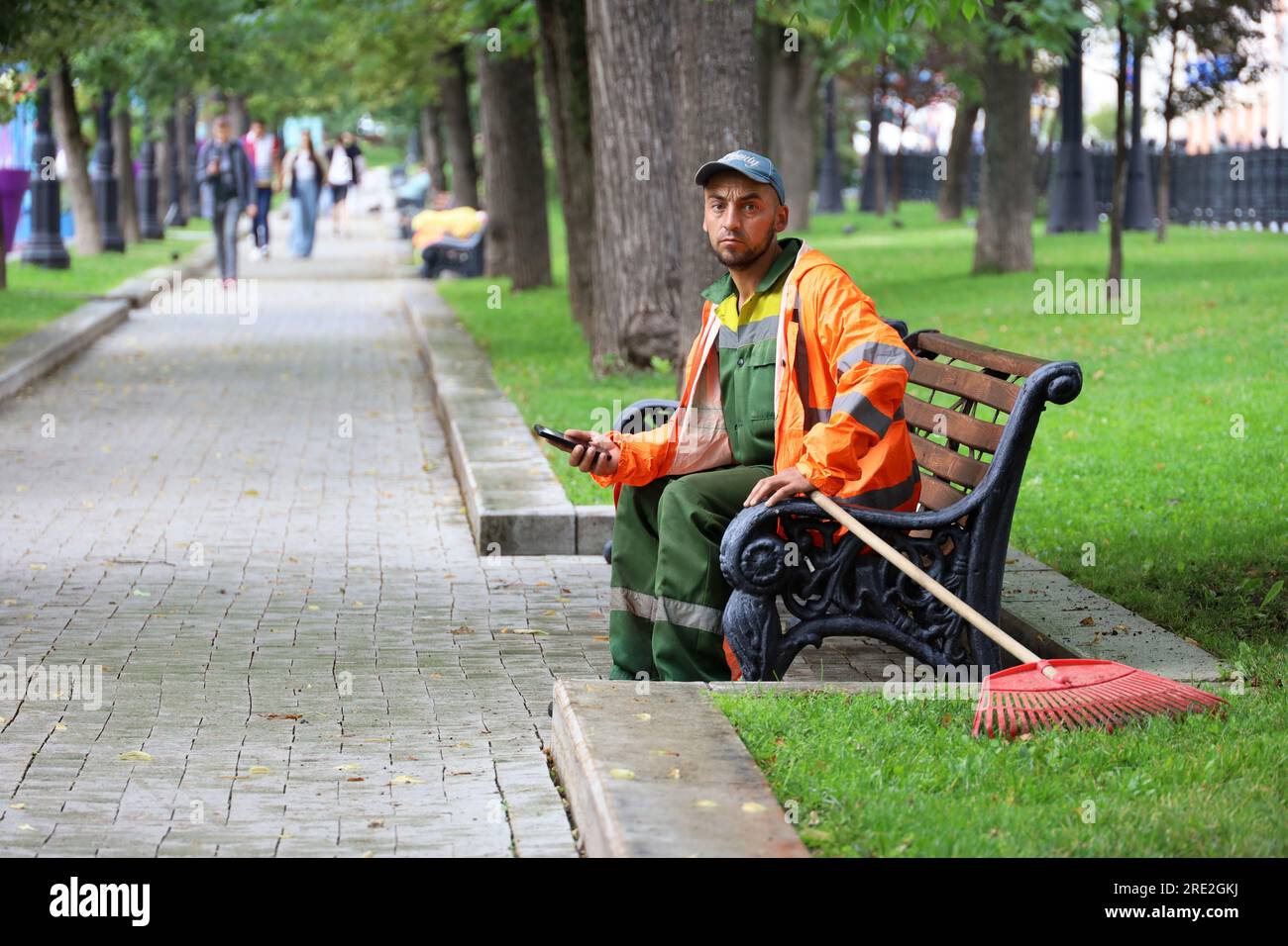 Cameriere uomo seduto con smartphone su una panchina nel parco estivo. Operaio dei servizi pubblici durante il riposo, pulizia delle strade Foto Stock