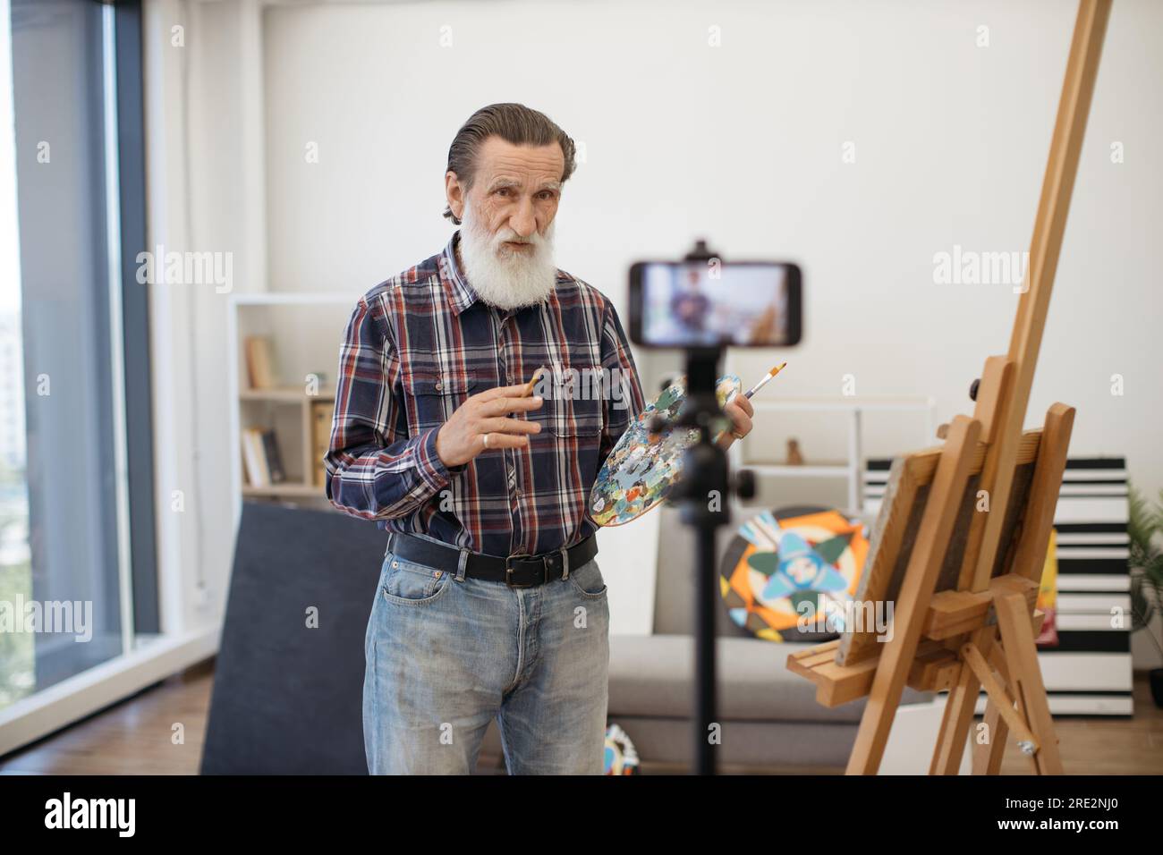 Gentiluomo anziano con lunga barba grigia in piedi davanti al treppiede con telefono cellulare e che parla delle tecniche di disegno in arte. Profes vestiti casualmente Foto Stock