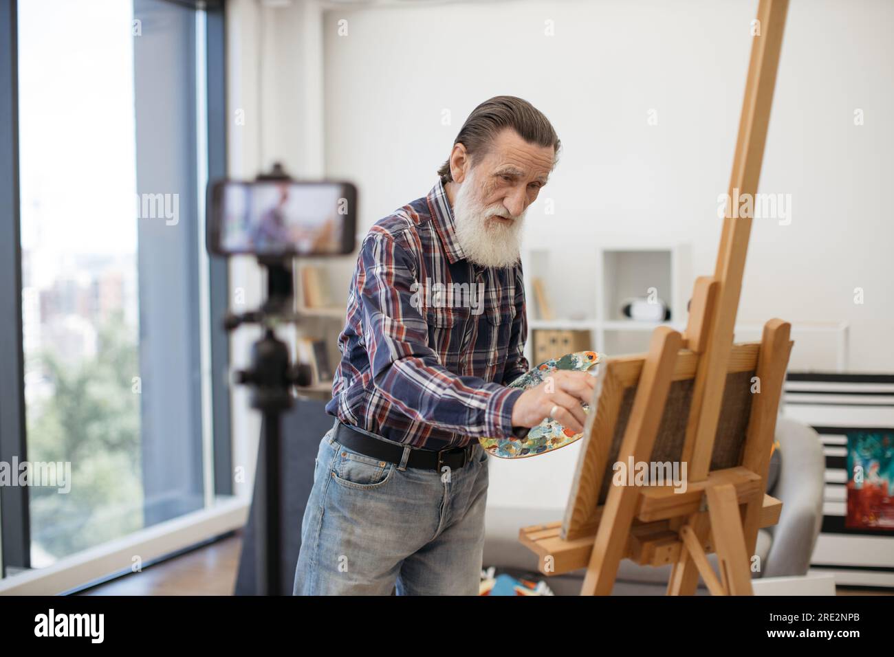 Gentiluomo anziano con lunga barba grigia in piedi davanti al treppiede con telefono cellulare e che parla delle tecniche di disegno in arte. Profes vestiti casualmente Foto Stock