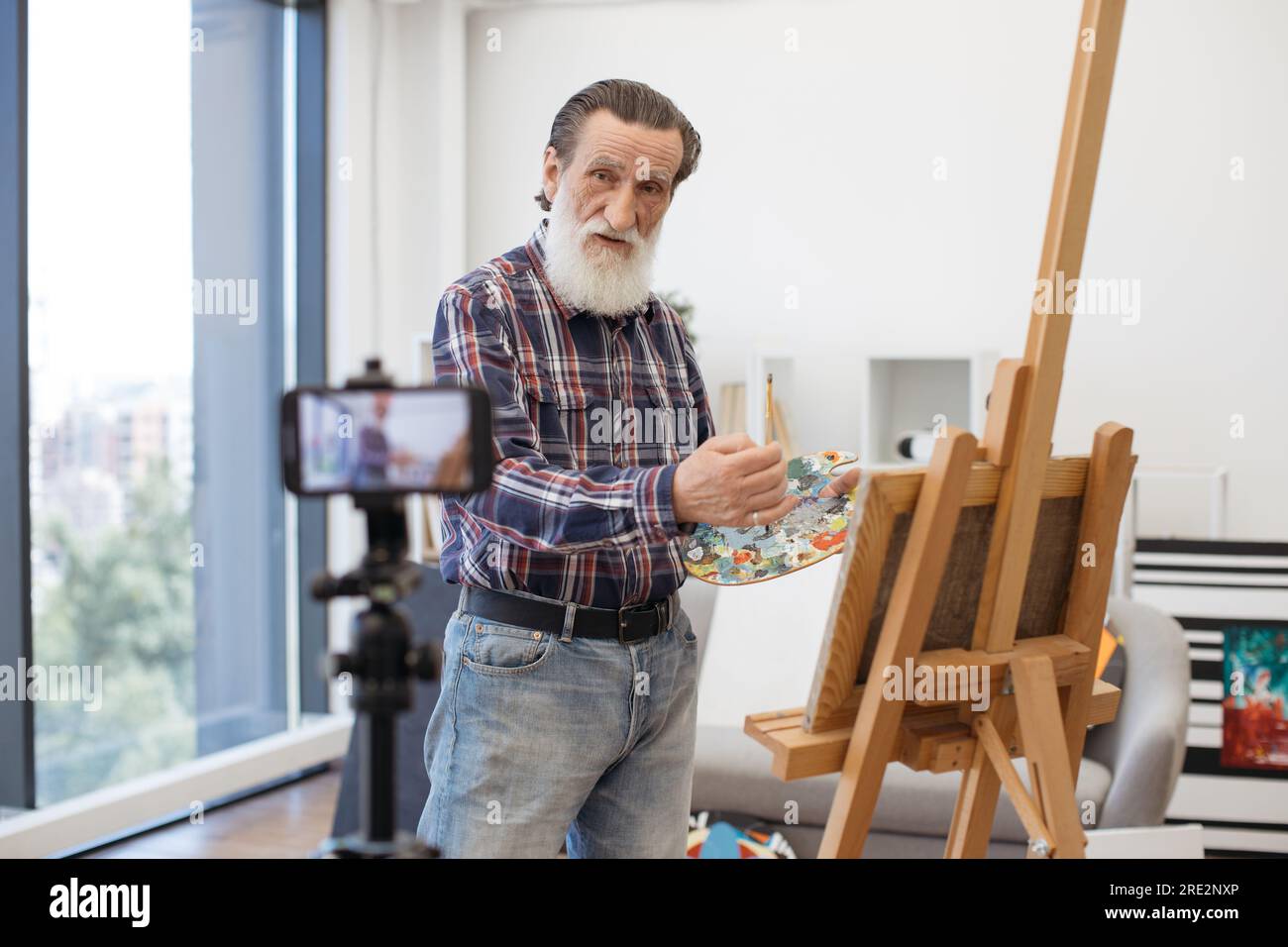 Gentiluomo anziano con lunga barba grigia in piedi davanti al treppiede con telefono cellulare e che parla delle tecniche di disegno in arte. Profes vestiti casualmente Foto Stock