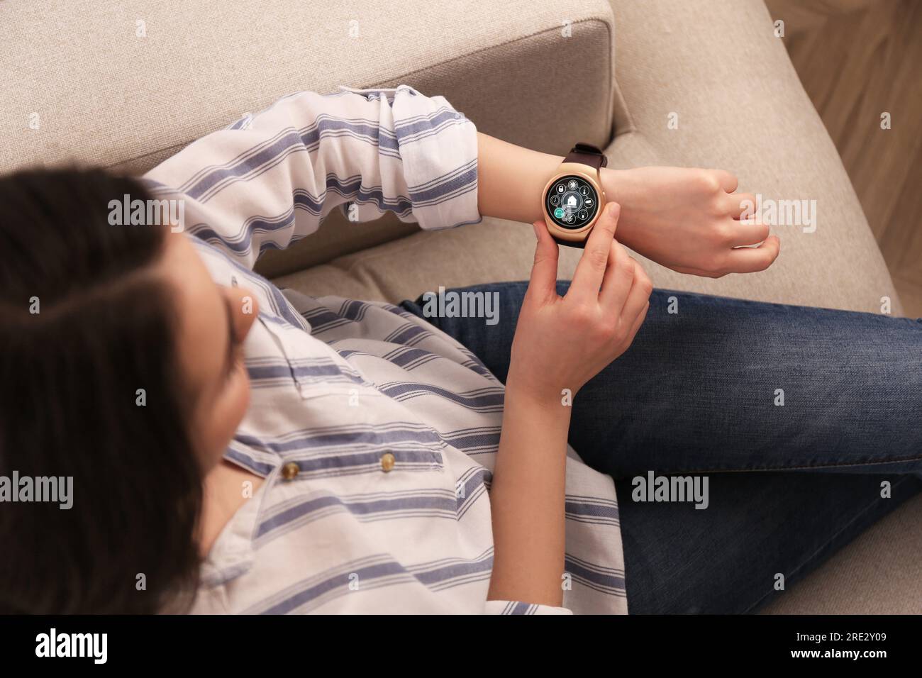 Donna che imposta il sistema di controllo della casa intelligente tramite smartwatch in interni, vista dall'alto. Interfaccia app con icone sul display Foto Stock