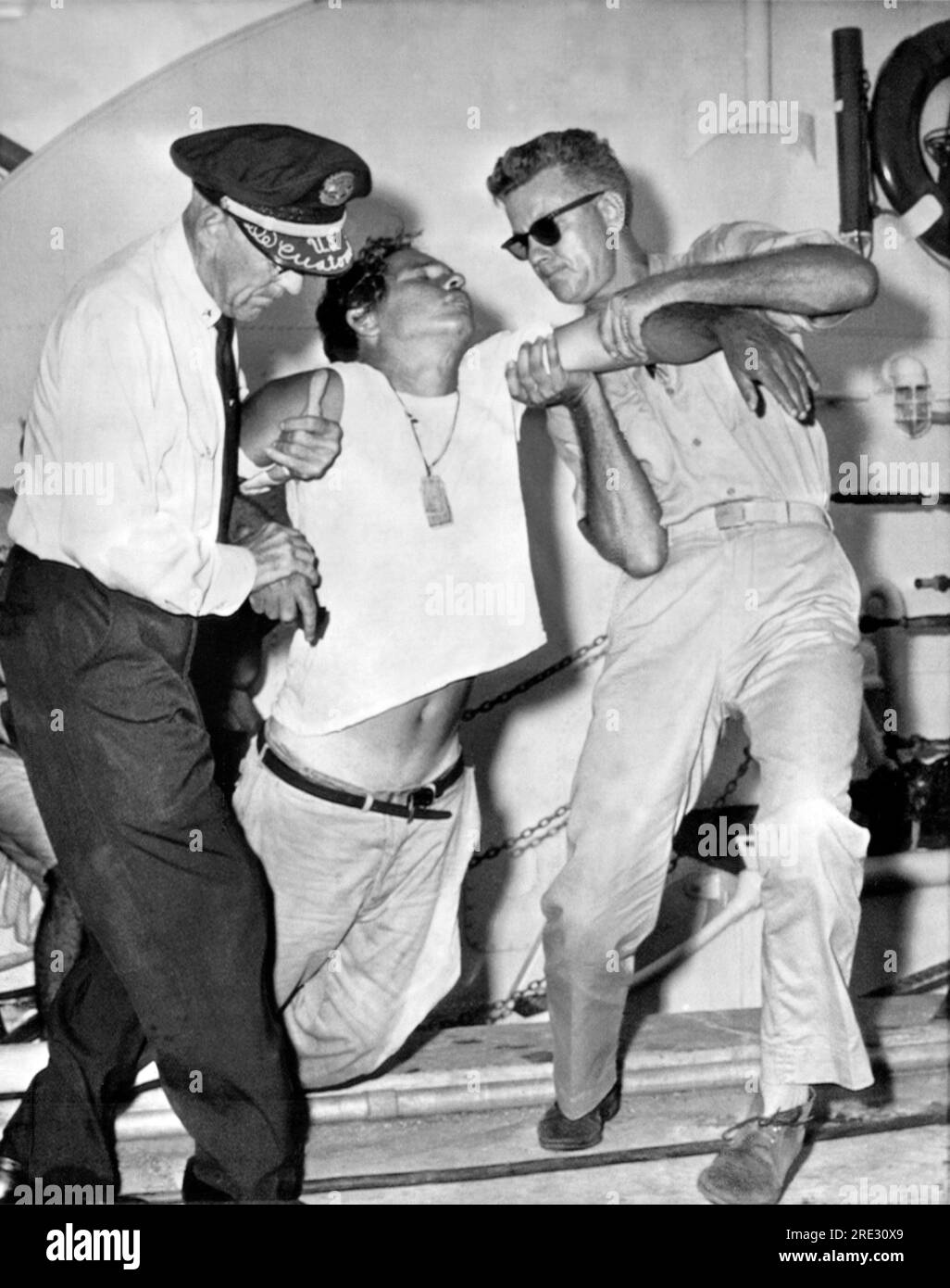 Miami, Florida: 16 agosto 1962 Un rifugiato cubano crolla mentre viene aiutato a terra da un U,S.. Un funzionario doganale e un U.S. Guardia costiera a Miami, Florida. Lui e altri cinque rifugiati erano stati alla deriva per almeno 8 giorni nella loro piccola barca a vela improvvisata, per lo più senza cibo o acqua. Foto Stock