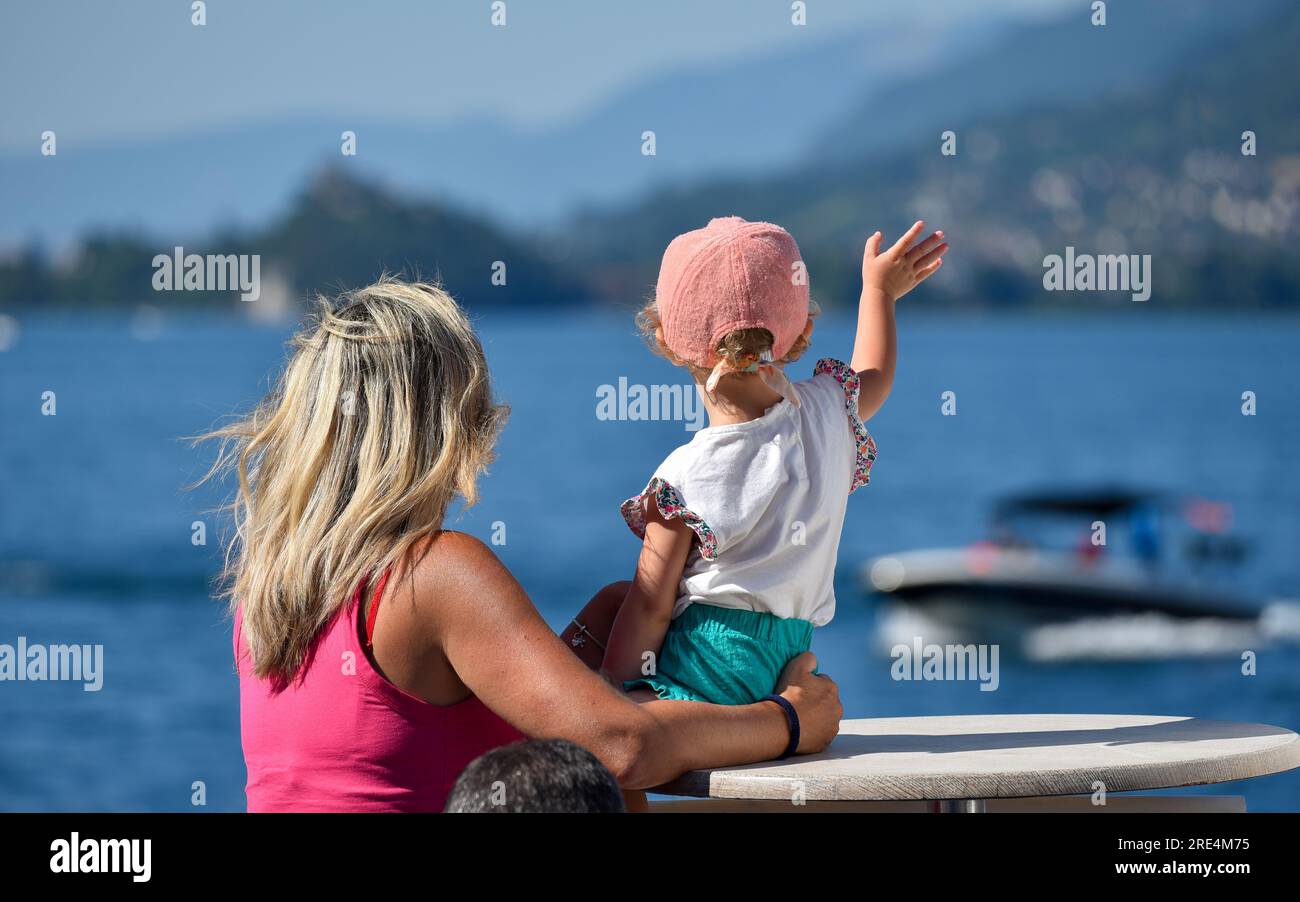 Una madre e suo figlio salutano una barca su un lago di montagna. Una madre bionda e sua figlia che indossa abiti rosa. Foto Stock