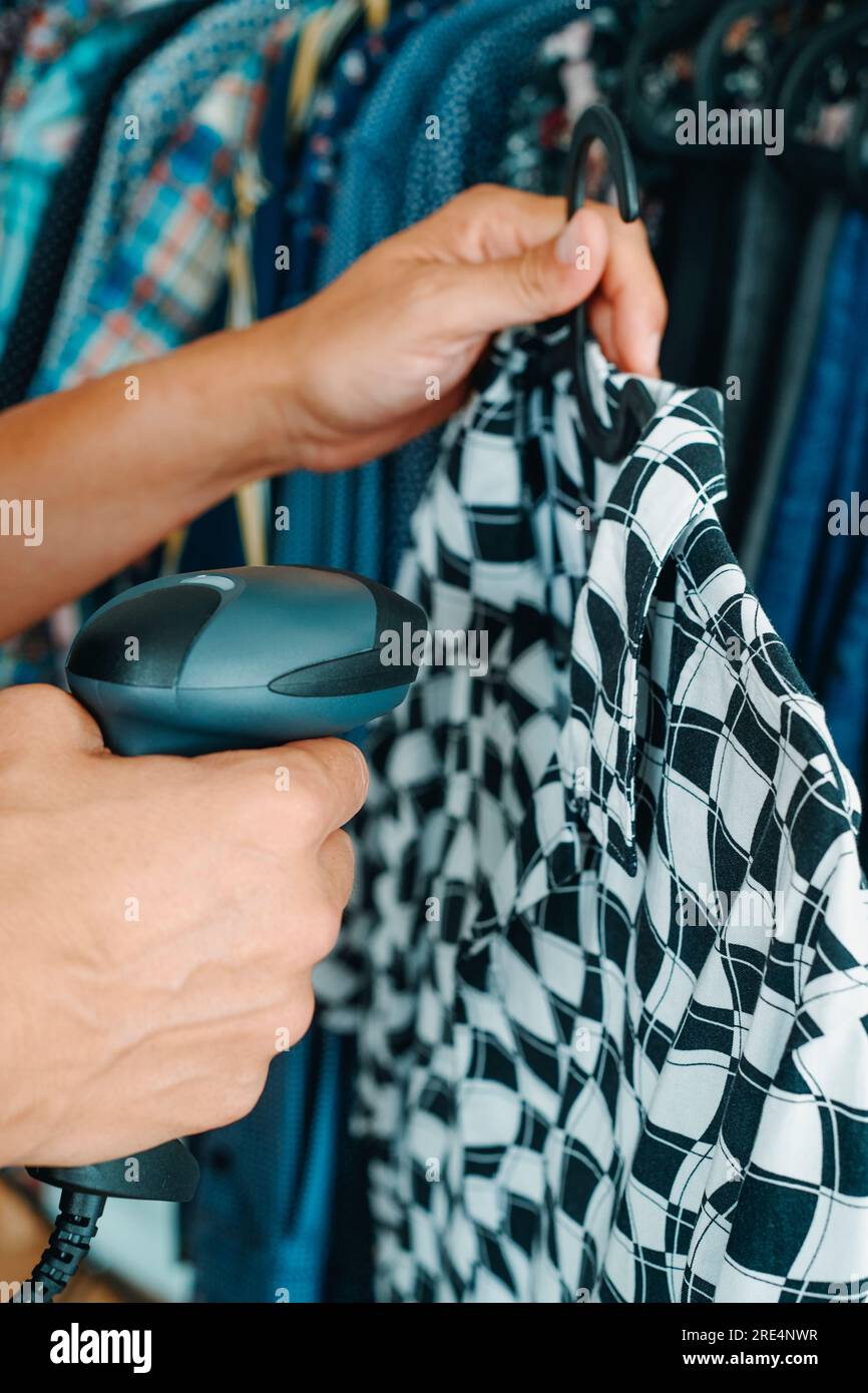 primo piano di un assistente in un negozio di abbigliamento che scansiona le informazioni di una camicia appesa a un appendiabiti utilizzando un lettore di codici a barre Foto Stock