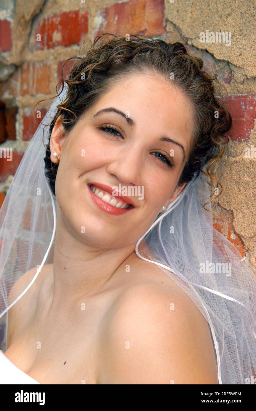 La sposa trasmittente si appoggia contro un muro di mattoni rustici. Lei è sorridente e felice. Indossa un abito e un velo senza spalline. Foto Stock