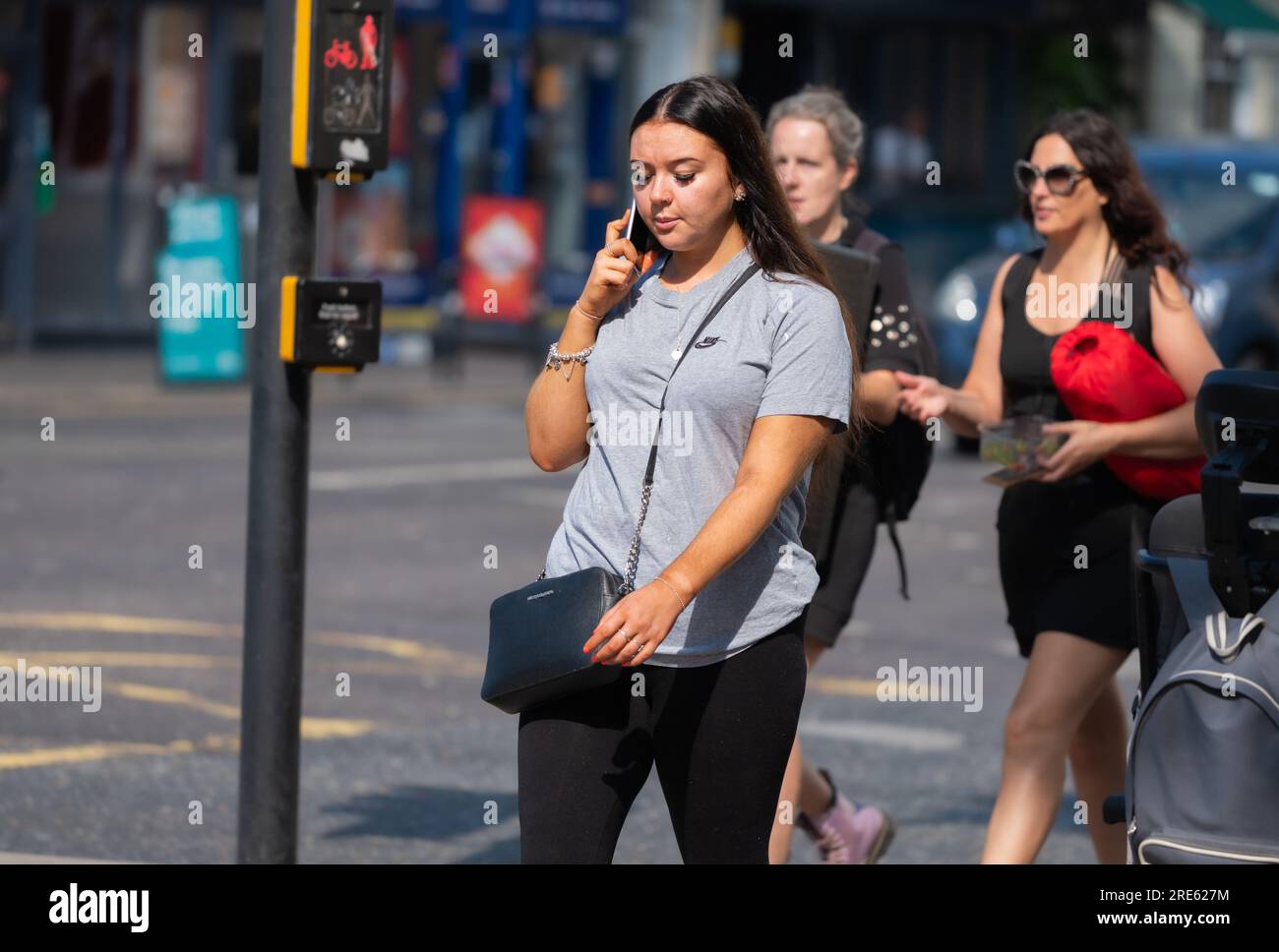 Giovane donna che cammina in una zona urbana trafficata e parla su un cellulare o cellulare, in estate in Inghilterra, Regno Unito. Foto Stock