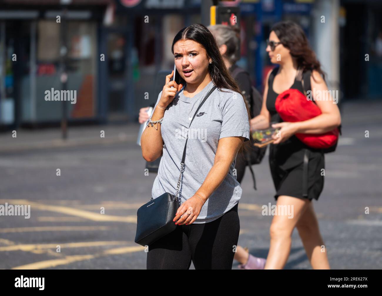 Giovane donna che cammina in una zona urbana trafficata e parla su un cellulare o cellulare, in estate in Inghilterra, Regno Unito. Foto Stock