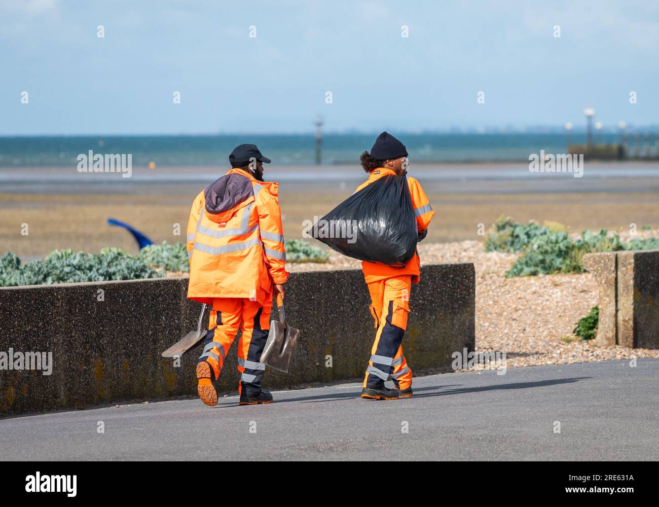 Un paio di lavoratori maschi, vestiti ad alta visibilità, passeggiate lungo la strada lungomare, raccolta di rifiuti dai bidoni della spazzatura e percorso di pulizia sul lungomare in estate, Regno Unito. Foto Stock