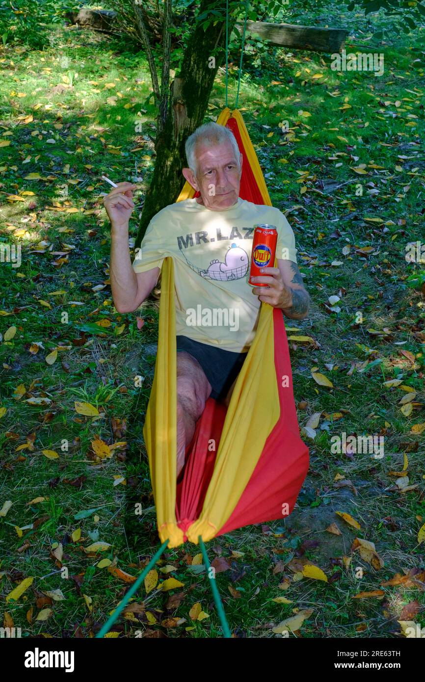 uomo anziano che si rilassa nell'amaca da giardino ombreggiata da alberi con sigaretta e lattina di birra indossando la maglietta mr. lazy t.shirt contea di zala ungheria Foto Stock