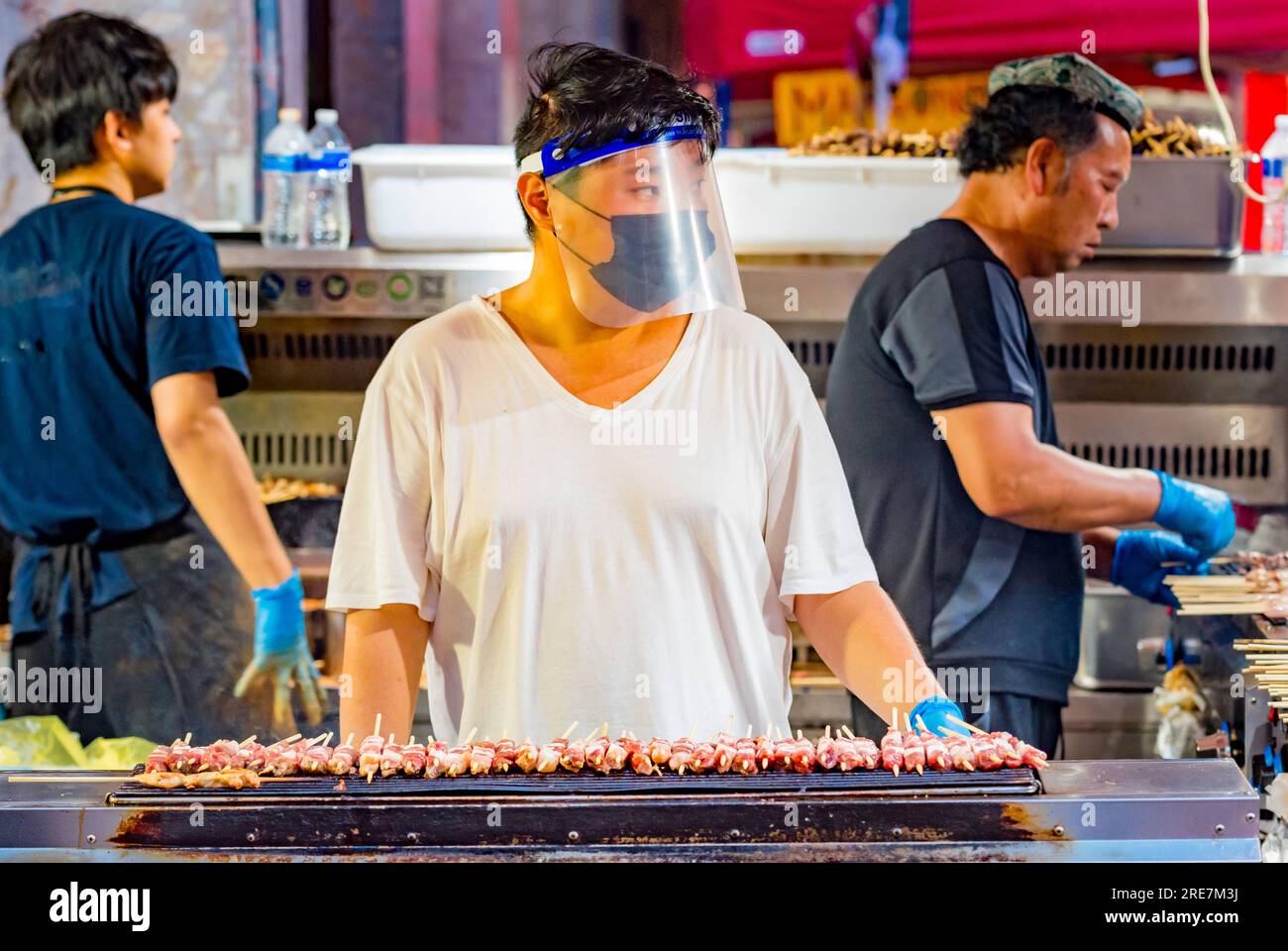 Una persona che indossa un panno e una maschera di protezione di plastica mentre cucina shishkebab in una bancarella di Street food a Sydney, Australia Foto Stock