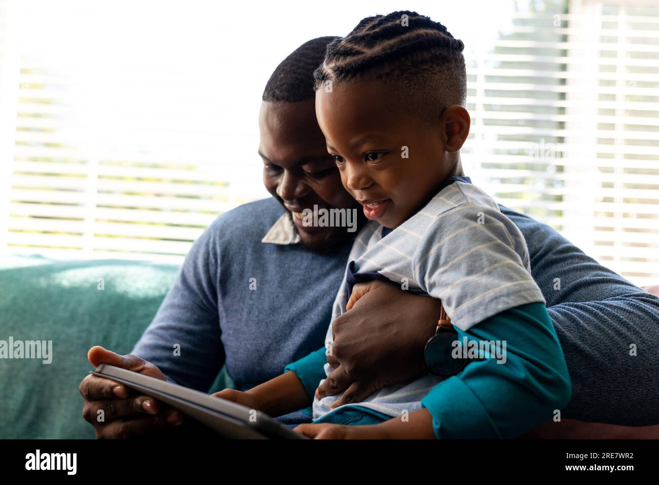Padre e figlio afroamericani felici seduti in salotto che abbracciano e usano un tablet Foto Stock