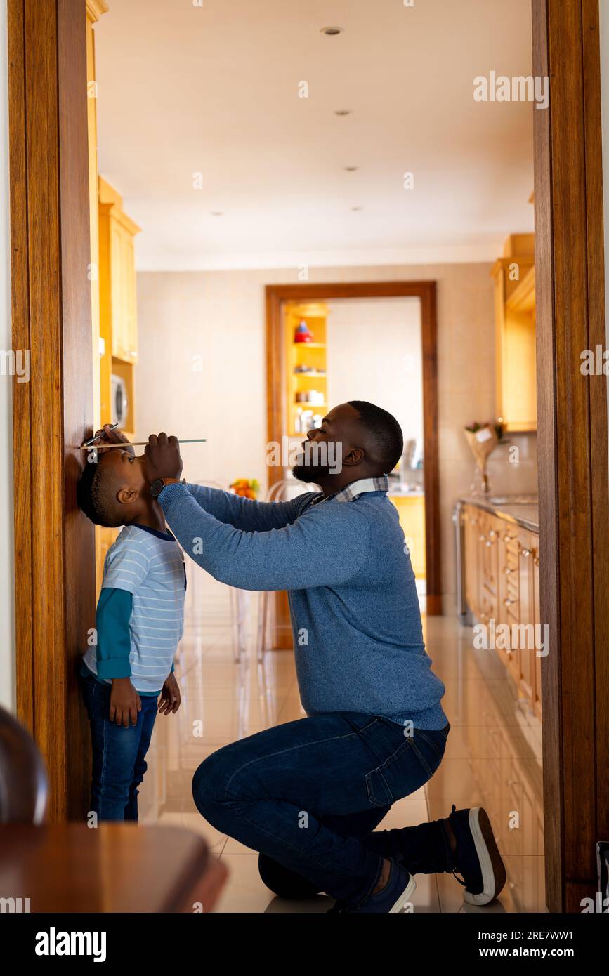 Padre afro-americano felice che misura il figlio e l'altezza di marcatura sul telaio della porta a casa, spazio copia Foto Stock