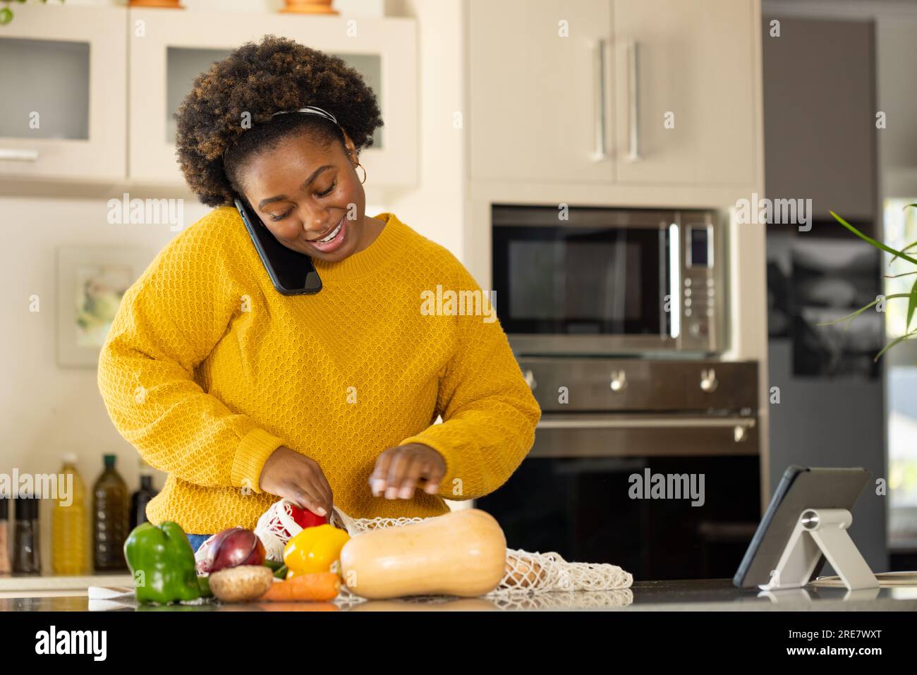 Una donna afroamericana felice e di taglia più grande che disimballa i generi alimentari e parla su smartphone in cucina Foto Stock