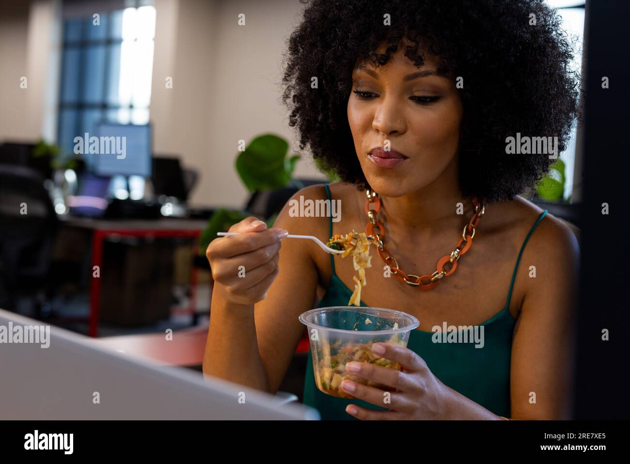 Donna afroamericana che fa uno spuntino seduto sulla sua scrivania in ufficio Foto Stock