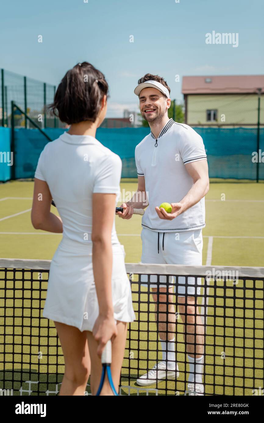 uomo felice che guarda la donna vicino alla rete da tennis, coppia in piedi sul campo da tennis, abbigliamento attivo, hobby Foto Stock
