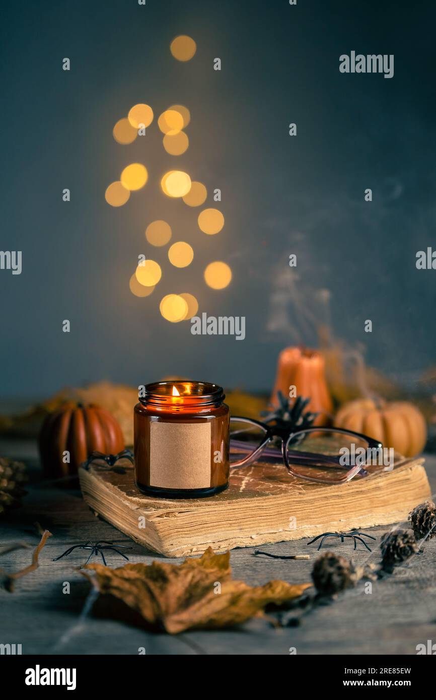 Candela Halloween STILL Life con cera di soia nel vaso di vetro ambrato scuro con etichetta vuota per il trucco. Candele a forma di zucca con coni, foglie, Old Book Foto Stock