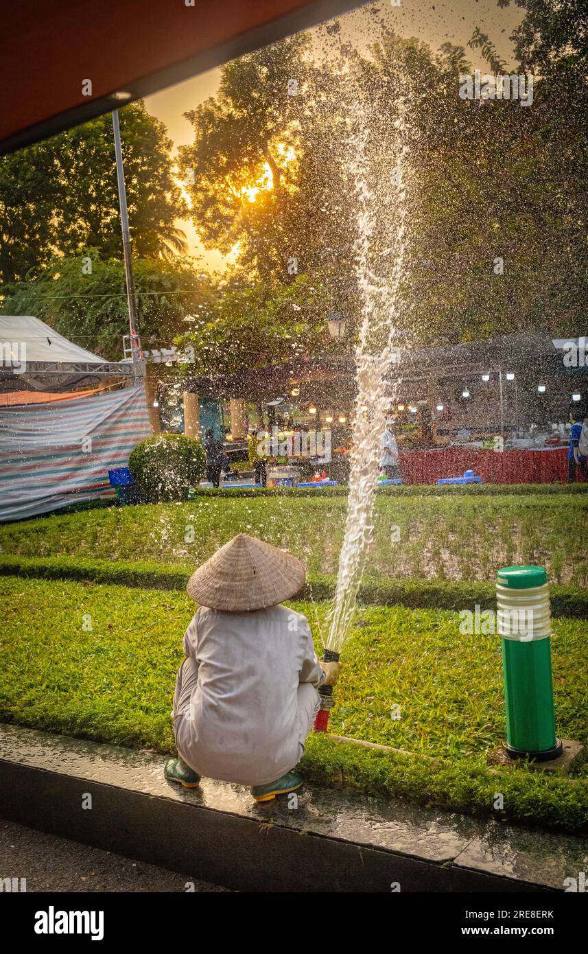 Una donna giardiniera vietnamita che indossa un cappello conico si accosta mentre spruzza acqua sulle aiuole del Reunification Park, Hanoi, Vietnam. Foto Stock