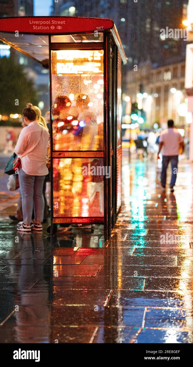 Persone che si riparano dalla pioggia di notte su una strada di Londra sotto un riparo degli autobus. Foto Stock