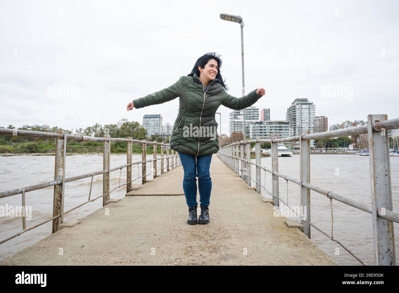 Giovane donna venezuelana in vacanza in Argentina felice di ballare in piedi sul molo nel porto di Olivos, indossa una giacca verde per il freddo Foto Stock