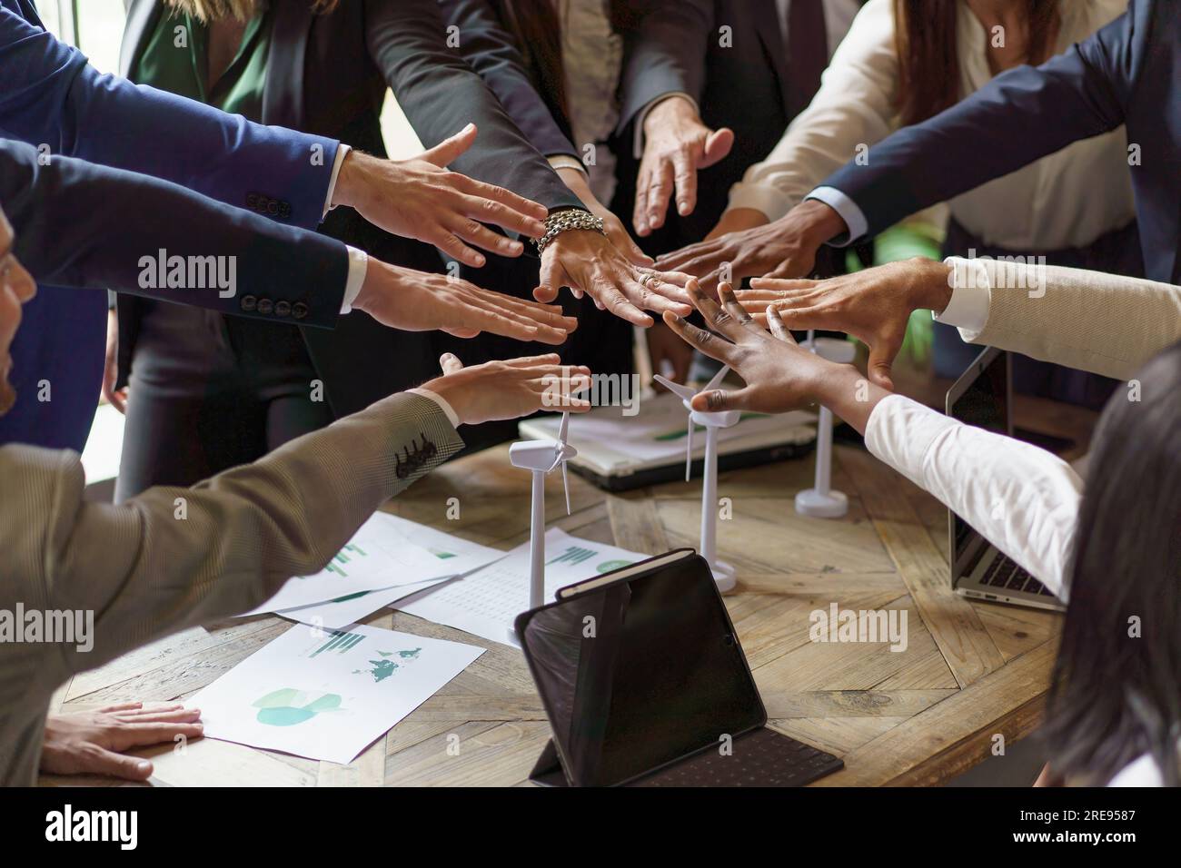 Il team multietnico raccoglie le mani sopra un tavolo disseminato di grafici e modelli di turbine eoliche, a significare vittoria e collaborazione in un'energia rinnovabile Foto Stock