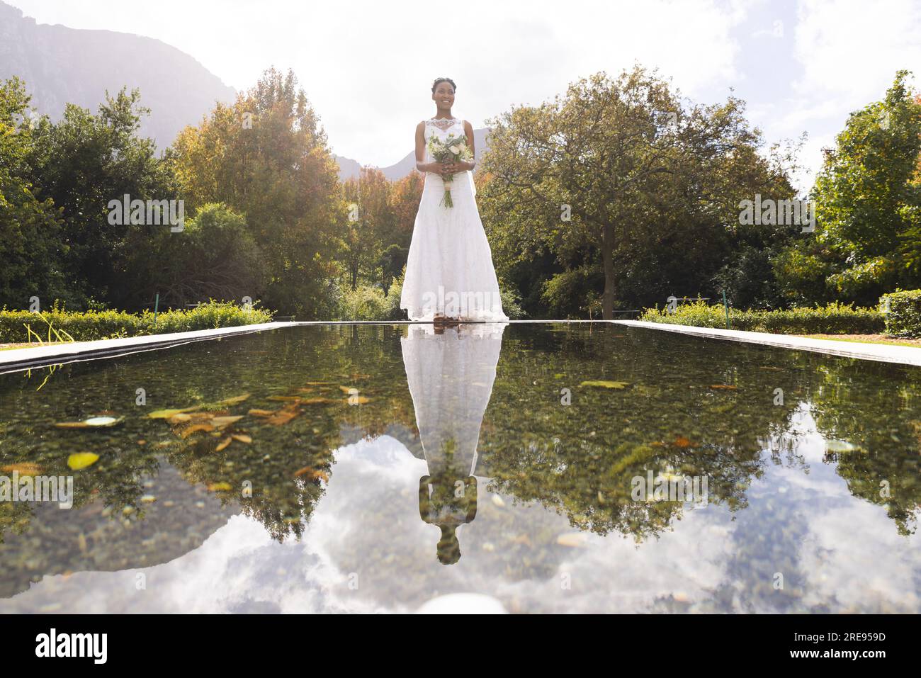 Felice sposa afro-americana con bouquet, riflesso nello stagno, in piedi in un giardino soleggiato Foto Stock