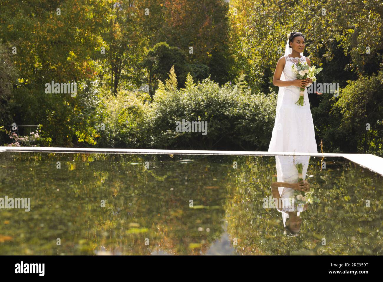 Felice sposa afro-americana con bouquet in piedi vicino allo stagno nel giardino soleggiato, spazio per le copie Foto Stock