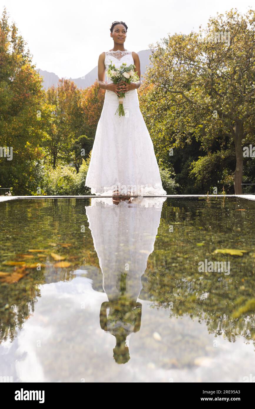 Felice sposa afro-americana con bouquet, riflesso nel laghetto, in un giardino soleggiato, spazio fotocopie Foto Stock