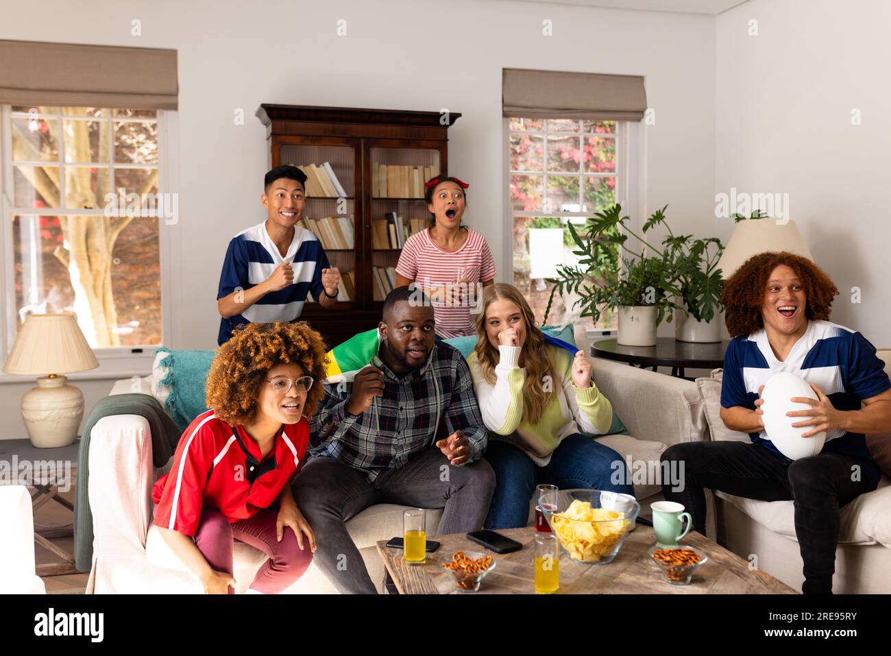 Felice gruppo di amici diversi con pallone da rugby e spuntini a casa Foto Stock