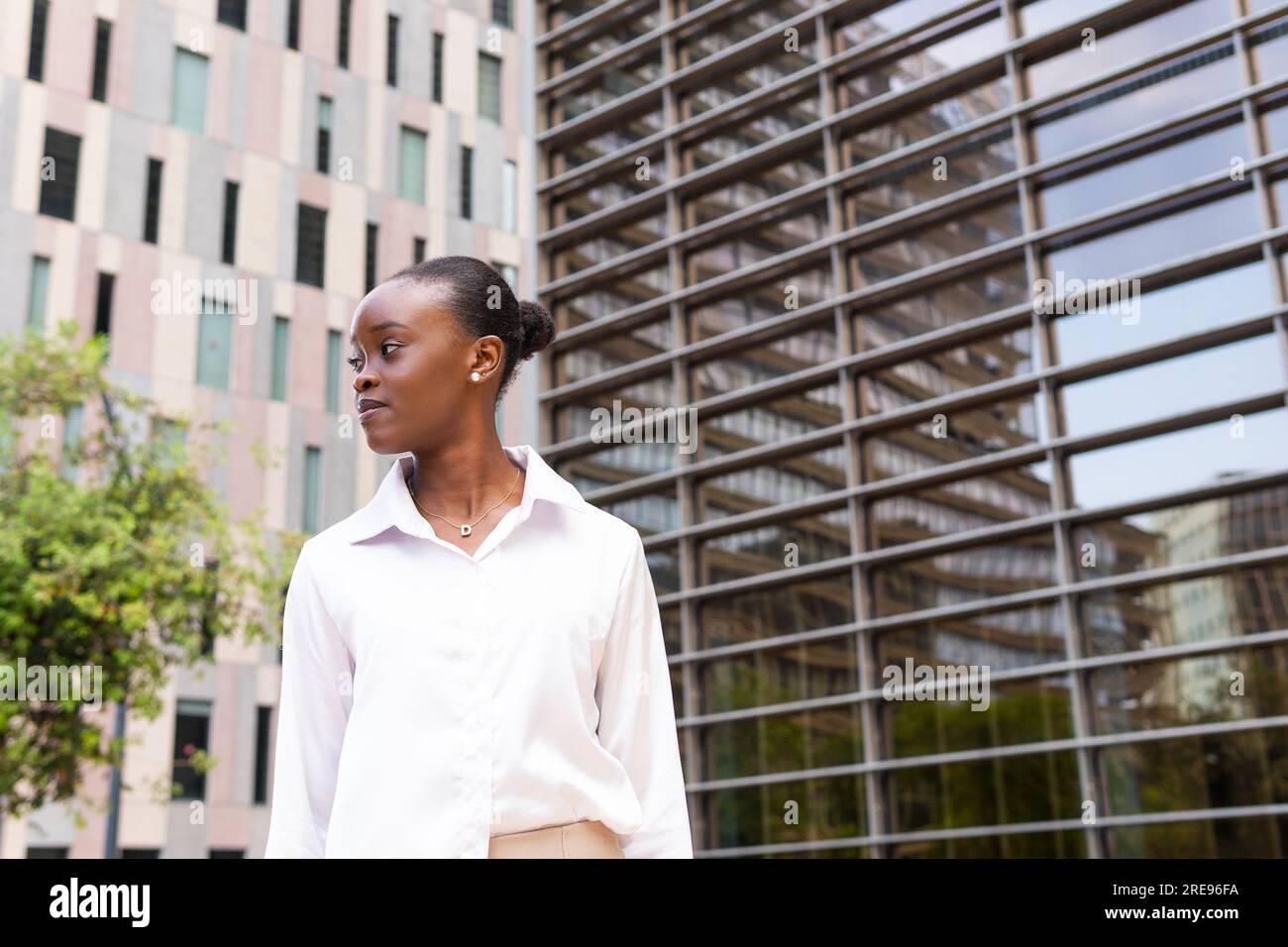 Vista laterale della giovane donna afroamericana in camicia bianca che guarda lontano mentre si trova vicino agli edifici moderni della città Foto Stock