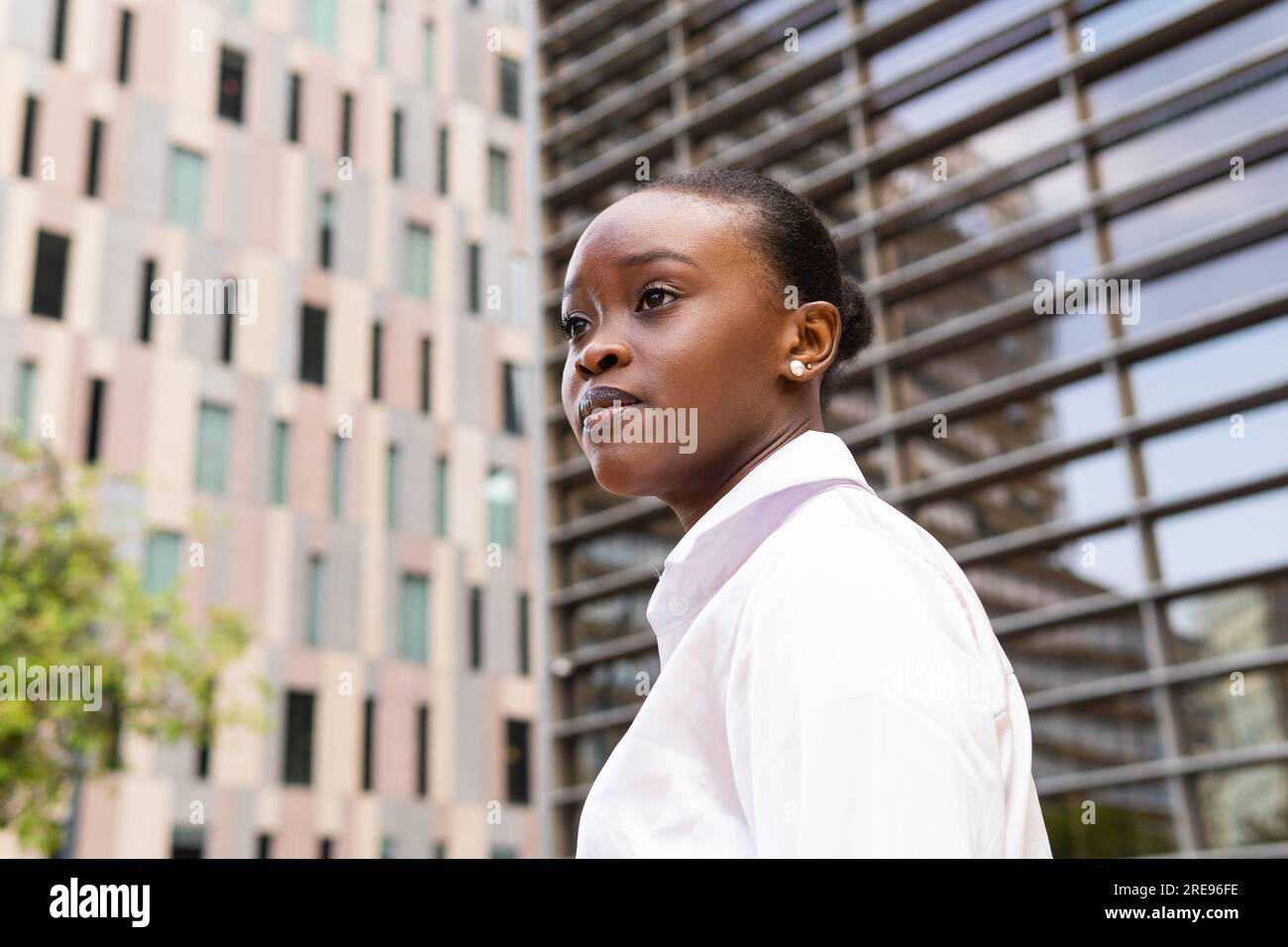 Dal basso, vista laterale della giovane donna afroamericana in camicia bianca che guarda lontano mentre si trova vicino agli edifici moderni della città Foto Stock
