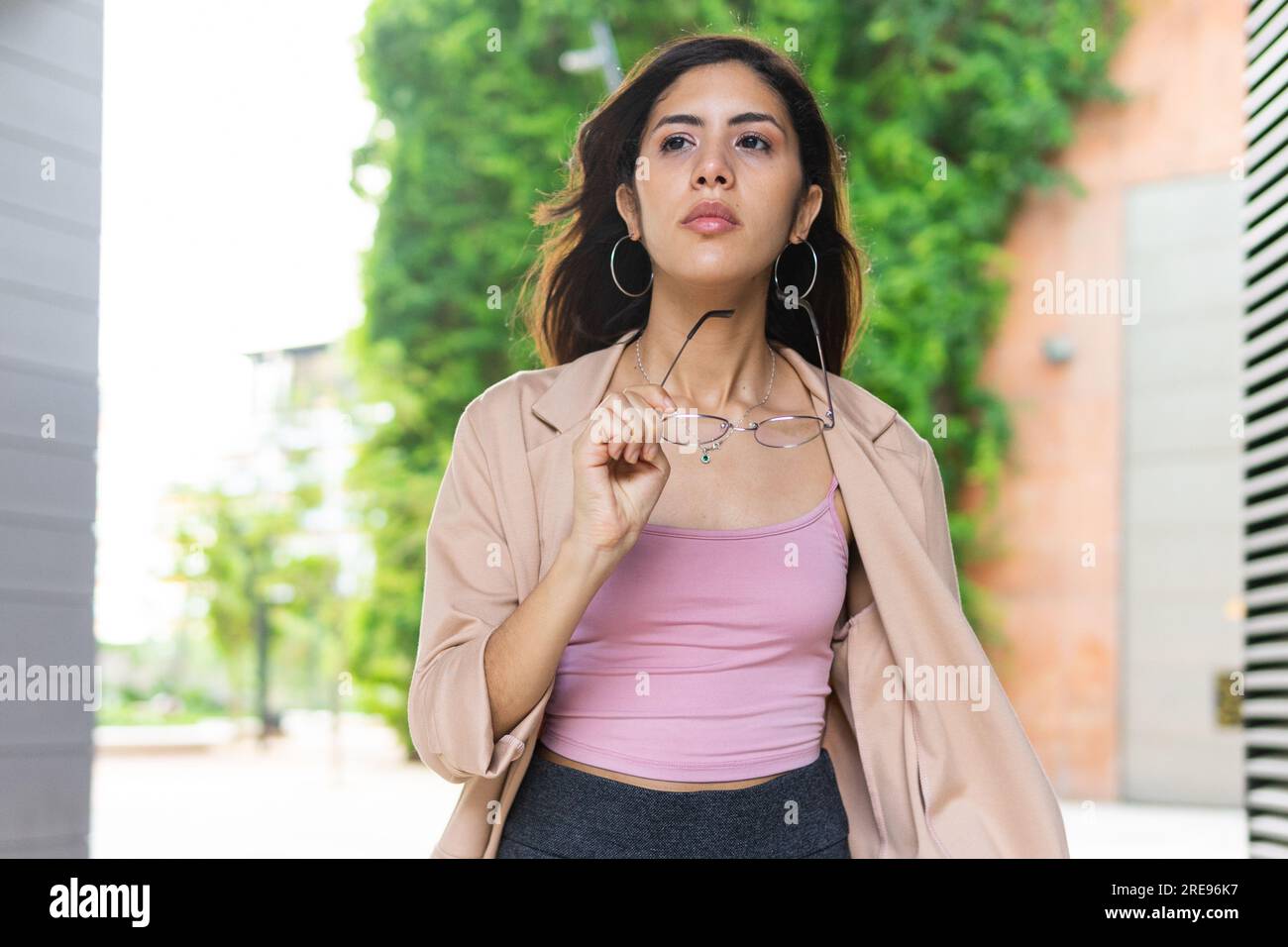Una giovane imprenditrice ispanica che cammina per strada urbana e regge occhiali da vista Foto Stock