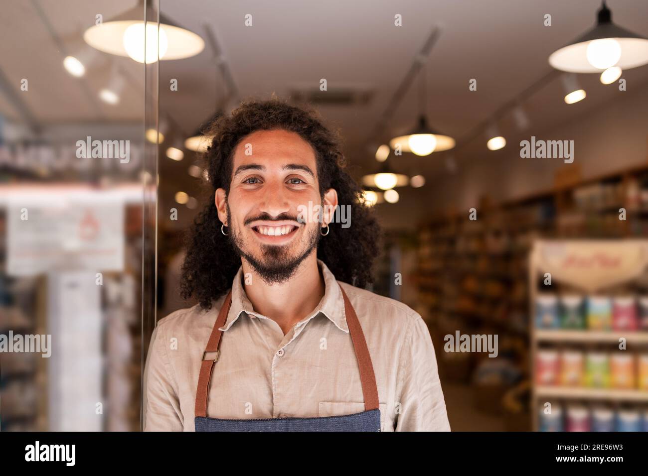 Ritratto di un uomo barbuto positivo in grembiule sorridente e che guarda la macchina fotografica mentre si trova vicino al buio sfondo illuminato sfocato nel supermercato Foto Stock