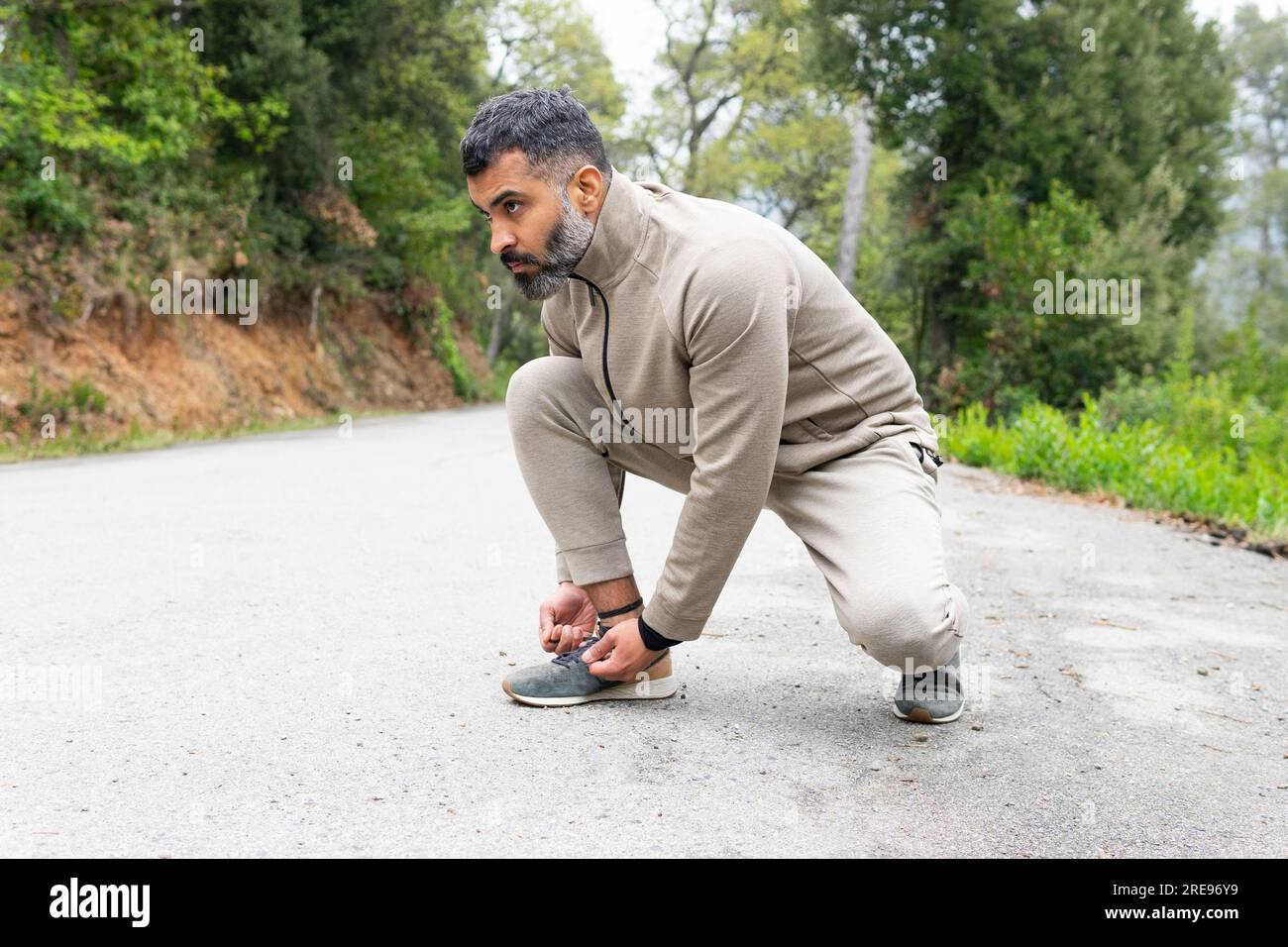 Vista laterale di un atleta indiano barbuto con abbigliamento sportivo che guarda lontano mentre si siede su asfalto e si lega un paio di scarpe da ginnastica vicino a alberi verdi Foto Stock