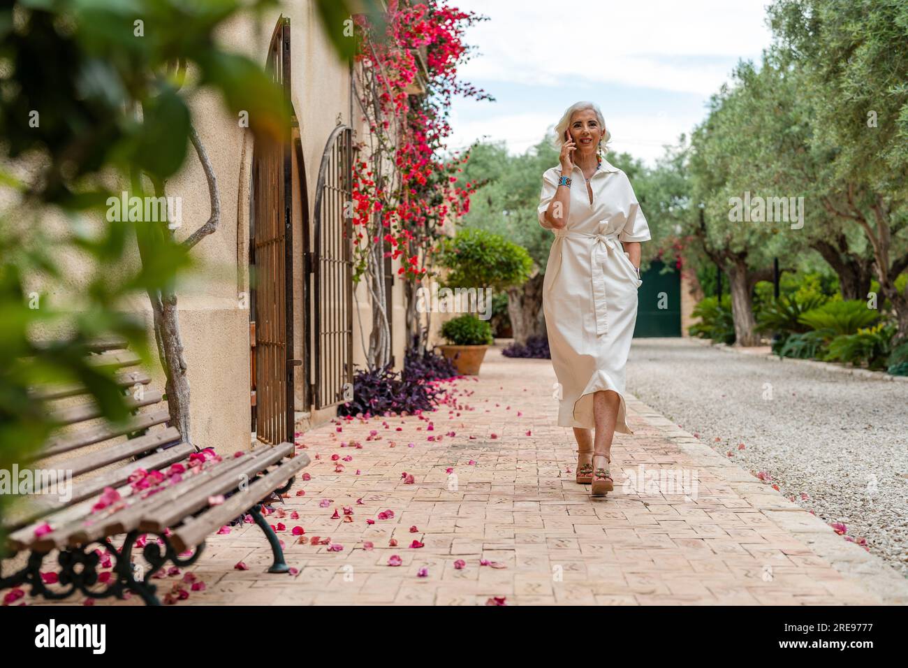 Donna anziana sorridente con i capelli grigi e con la mano nella tasca che guarda la fotocamera mentre cammina sul marciapiede con la panchina delle piante con porte di recinzione Foto Stock
