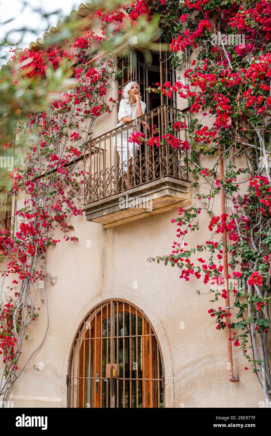 Angolo basso di donna anziana dai capelli grigi che guarda lontano mentre si sta in piedi con le mani su un balcone recintato di ferro dell'edificio con fiori in fiore e leav verde Foto Stock