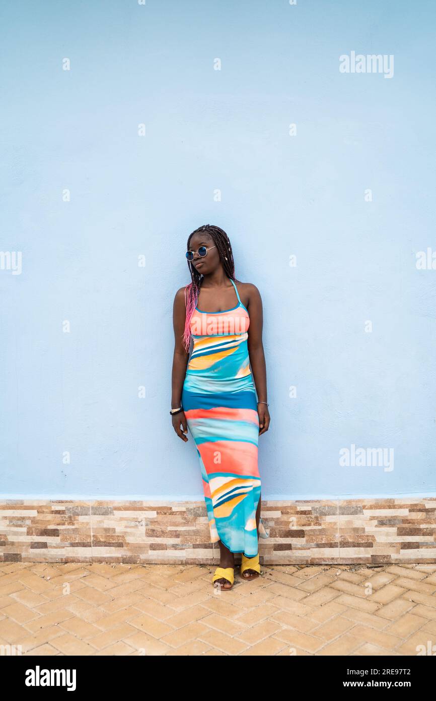 Corpo completo di giovane donna afro-americana in elegante abito colorato e occhiali da sole in piedi contro la moderna parete blu della casa mentre si guarda lontano Foto Stock