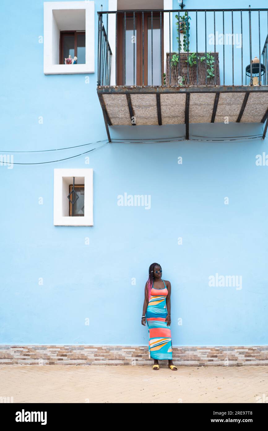 Corpo completo di giovane donna afro-americana in elegante abito colorato e occhiali da sole in piedi contro la moderna parete blu della casa mentre si guarda lontano Foto Stock