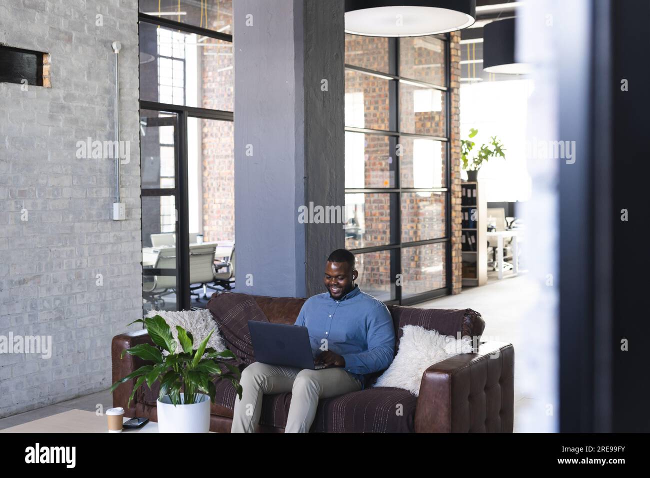 Felice uomo d'affari afroamericano informale seduto sul divano e che usa il computer portatile in ufficio Foto Stock