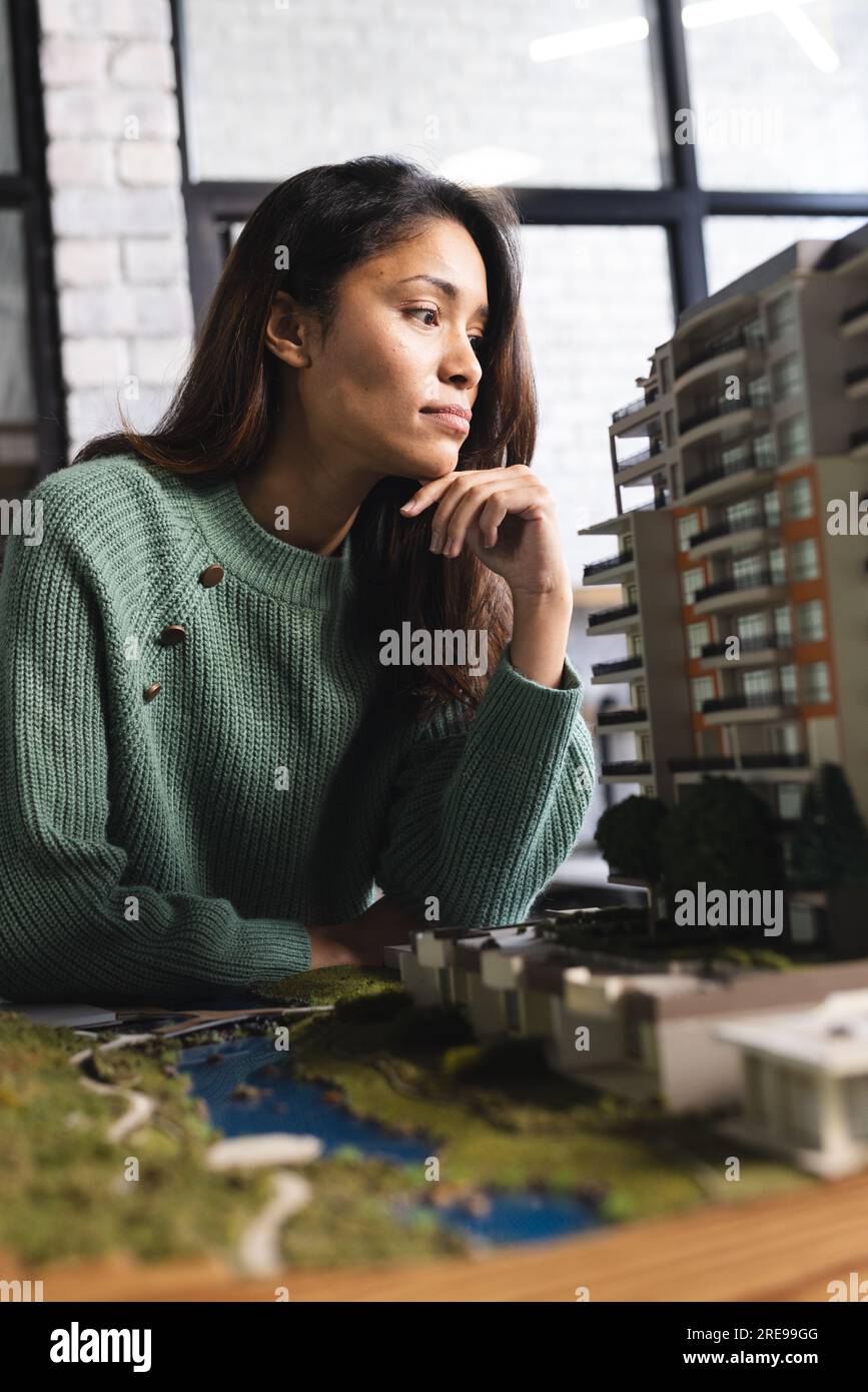Donna d'affari birazziale e concentrata che ispeziona il modello di edificio in ufficio Foto Stock