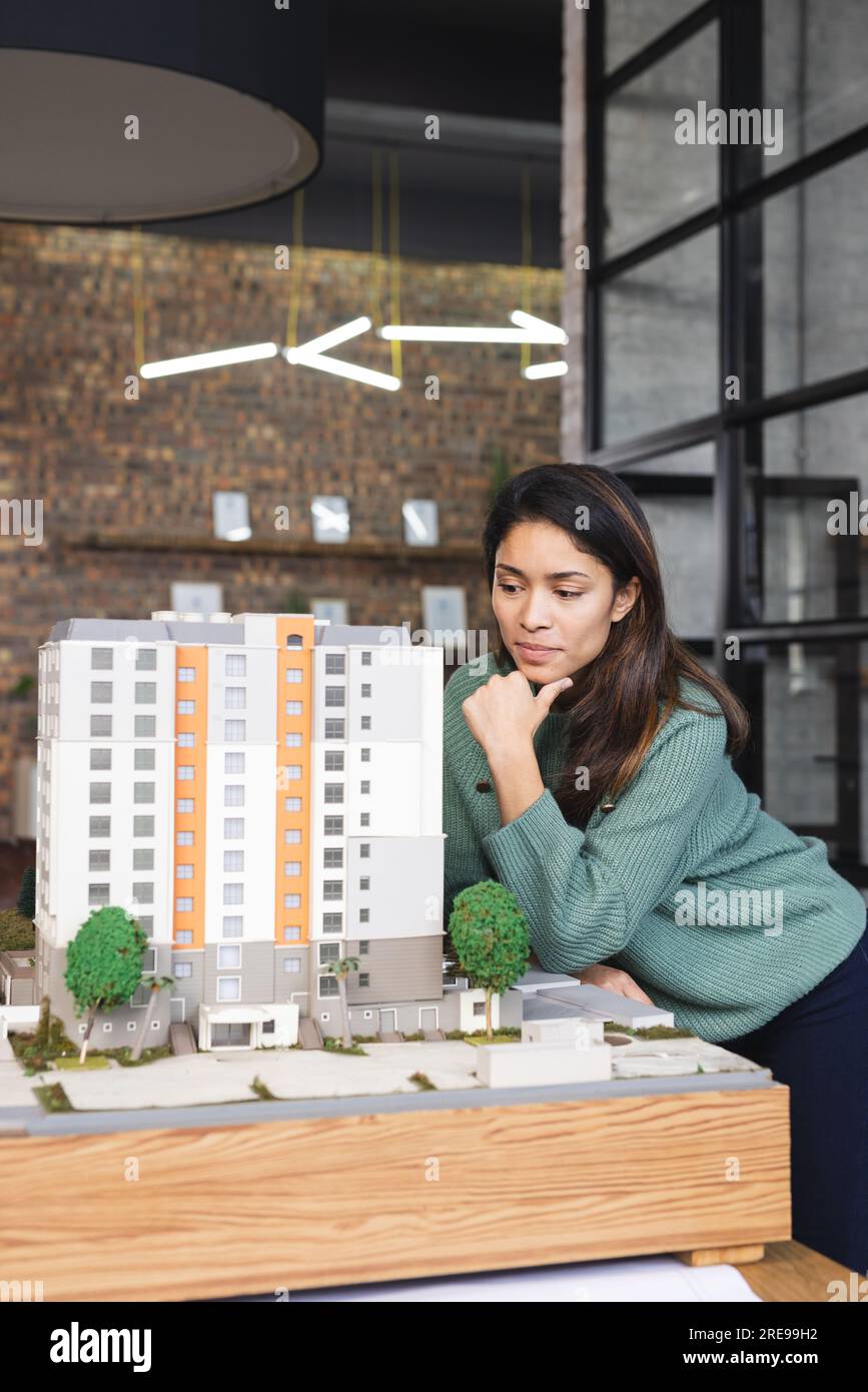 Donna d'affari birazziale e concentrata che ispeziona il modello di edificio in ufficio Foto Stock