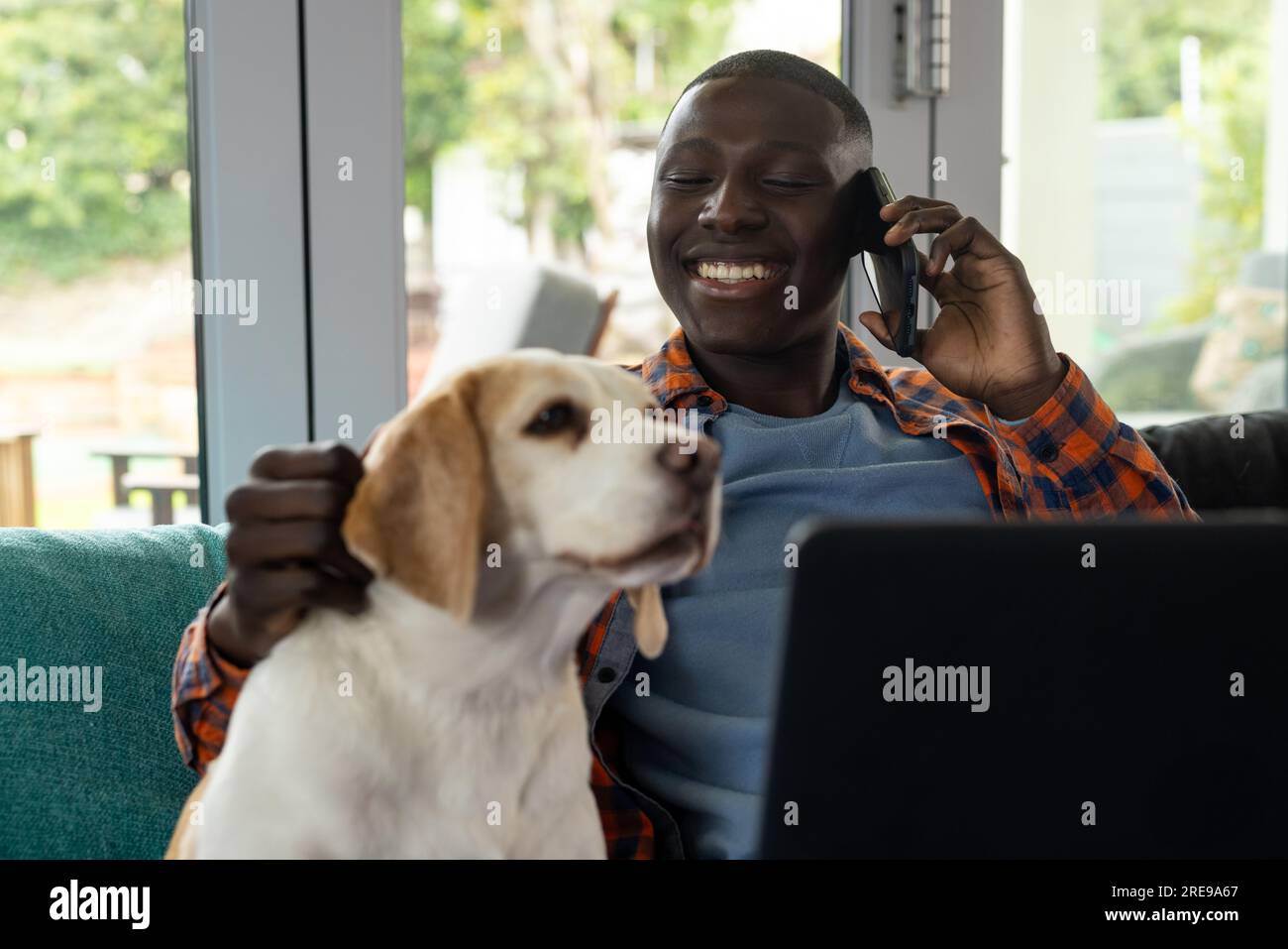 Uomo afro-americano seduto sul divano, che usa il computer portatile e parla sullo smartphone, con cane domestico a casa Foto Stock