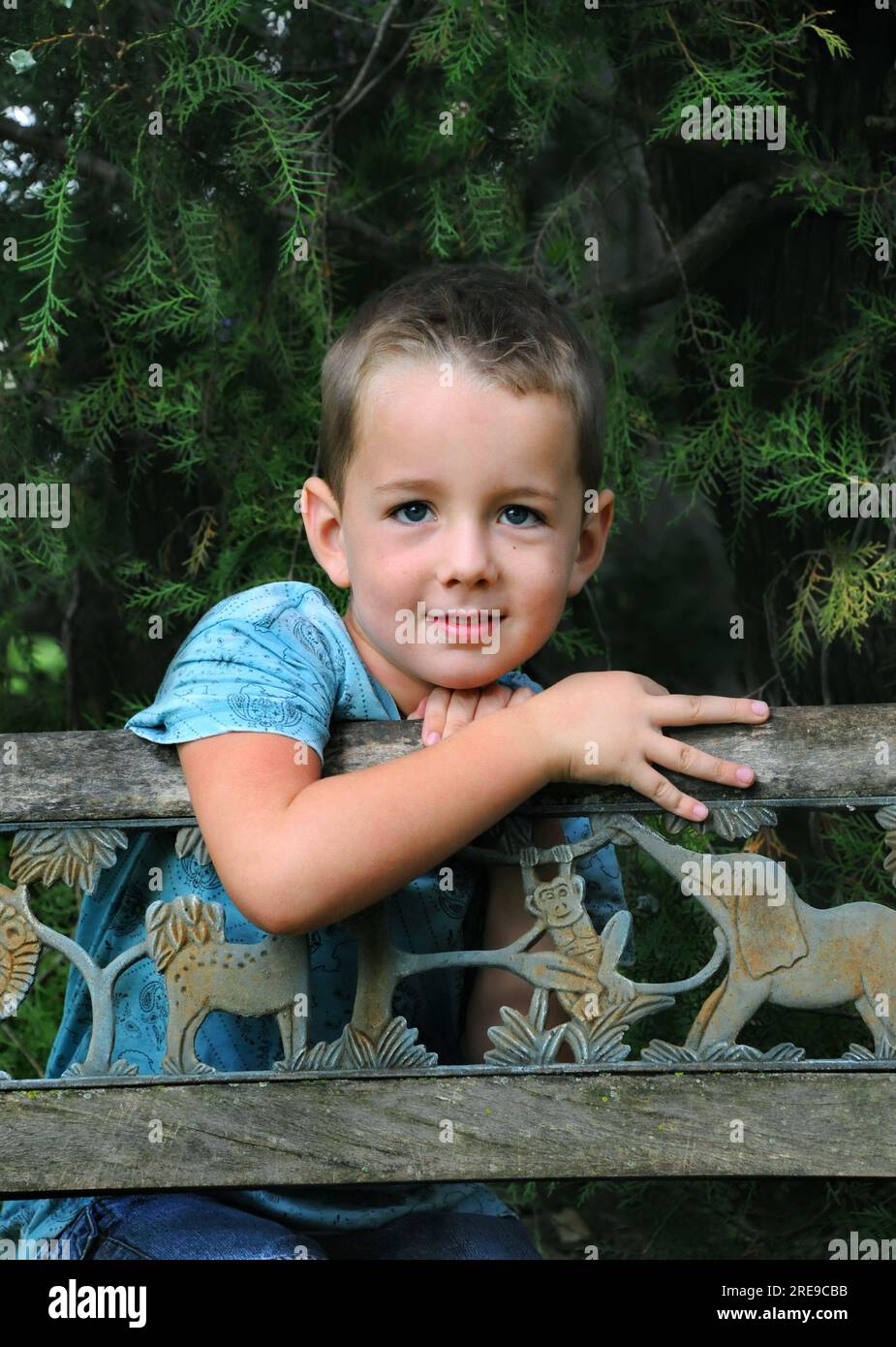 Il bambino si diverte a sognare un giorno da ragazzo, appoggiandosi su una panchina di legno in un parco alberato. Ha una camicia blu e dei jeans. Foto Stock