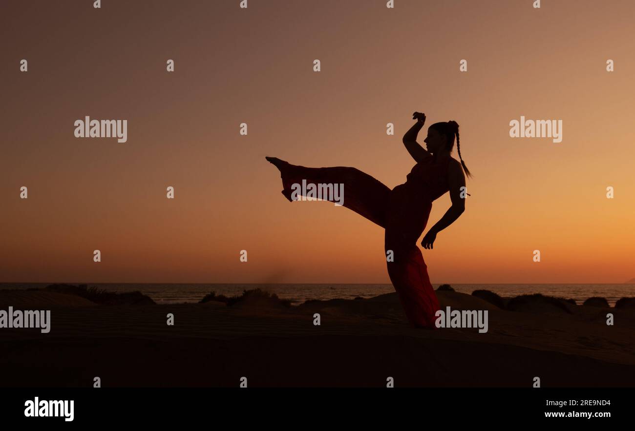 Silhouette di una donna che balla su una spiaggia sabbiosa al tramonto. Donna caucasica che posa all'aperto sulla costa. Foto Stock