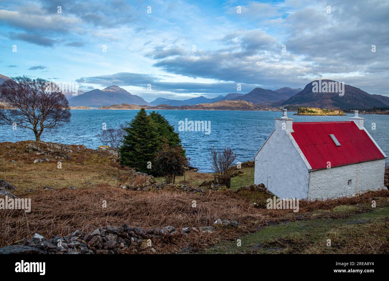 Caratteristico cottage con tetto in latta rossa ai margini di un loch affacciato sulle montagne di Torridon, Scozia Foto Stock