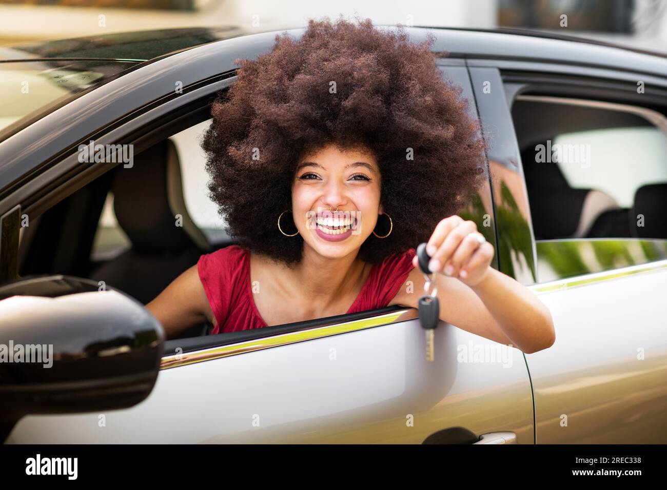 Felice donna afro-americana con capelli afro e trucco sorridente e guardando la macchina fotografica mentre si siede in una macchina nuova e mostra la chiave della macchina Foto Stock
