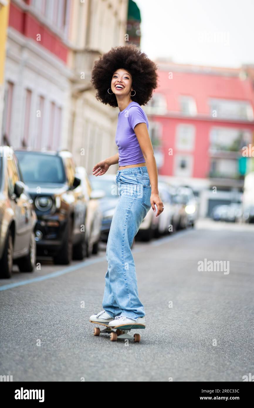 Vista laterale completa della vivace pattinatrice afro-americana in abiti casual con skateboard per equitazione afro su asfalto in città e in loo Foto Stock