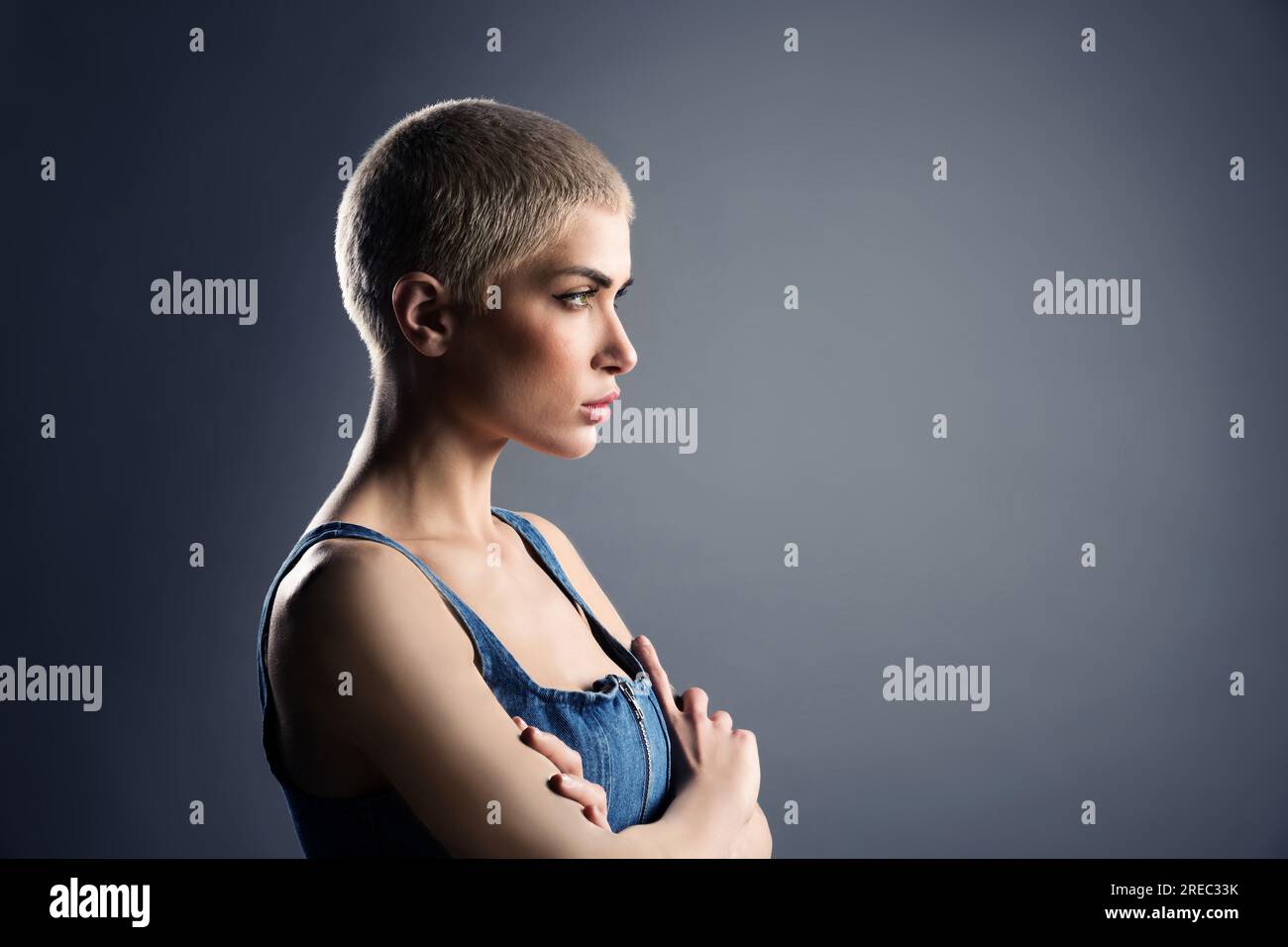 Vista laterale di una giovane modella donna seria con capelli biondi corti braccia incrociate e distanti dallo sguardo su sfondo grigio Foto Stock