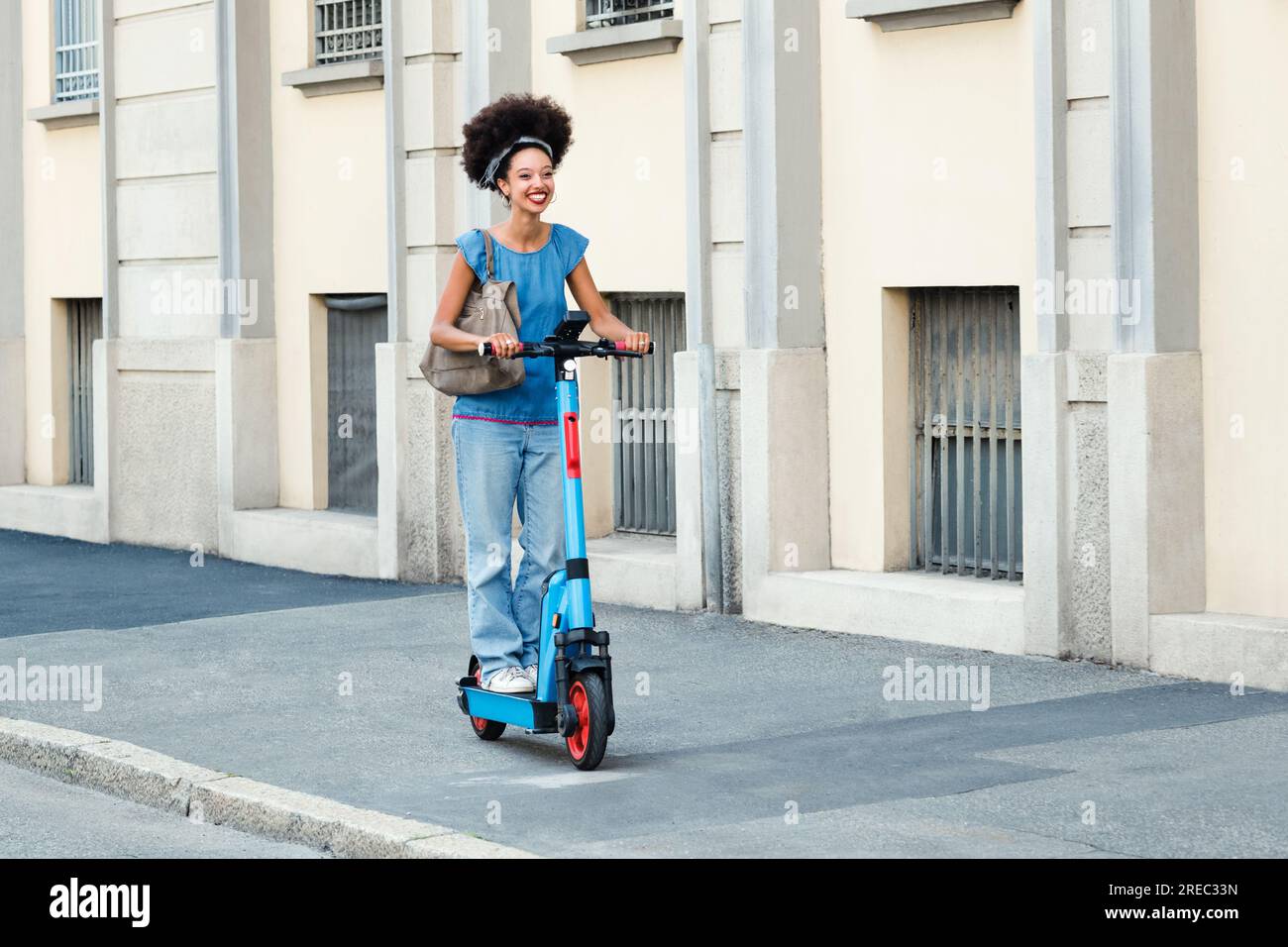 Corpo pieno di allegra giovane donna nera con acconciatura afro che guida uno scooter elettrico sulla strada della città alla luce del sole Foto Stock